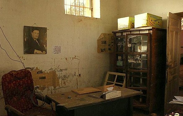 تظهر الصورة غرفة مهجورة مع جدران تحمل آثار الزمن، حيث تتواجد أريكة حمراء تقليدية على الجانبين. في الزاوية، يوجد مكتب خشبي بسيط بجانبه بعض الأوراق. على الجدار، توجد صورة معلقة لرجل يرتدي بدلة، مما يشير إلى أنه كان شخصية مهمة في الماضي. في الخلفية، يوجد زوج من خزائن الزجاج التي تحتوي على أشياء غير محددة. الإضاءة في الغرفة خافتة، مما يضيف إلى الأجواء الكئيبة للمكان.