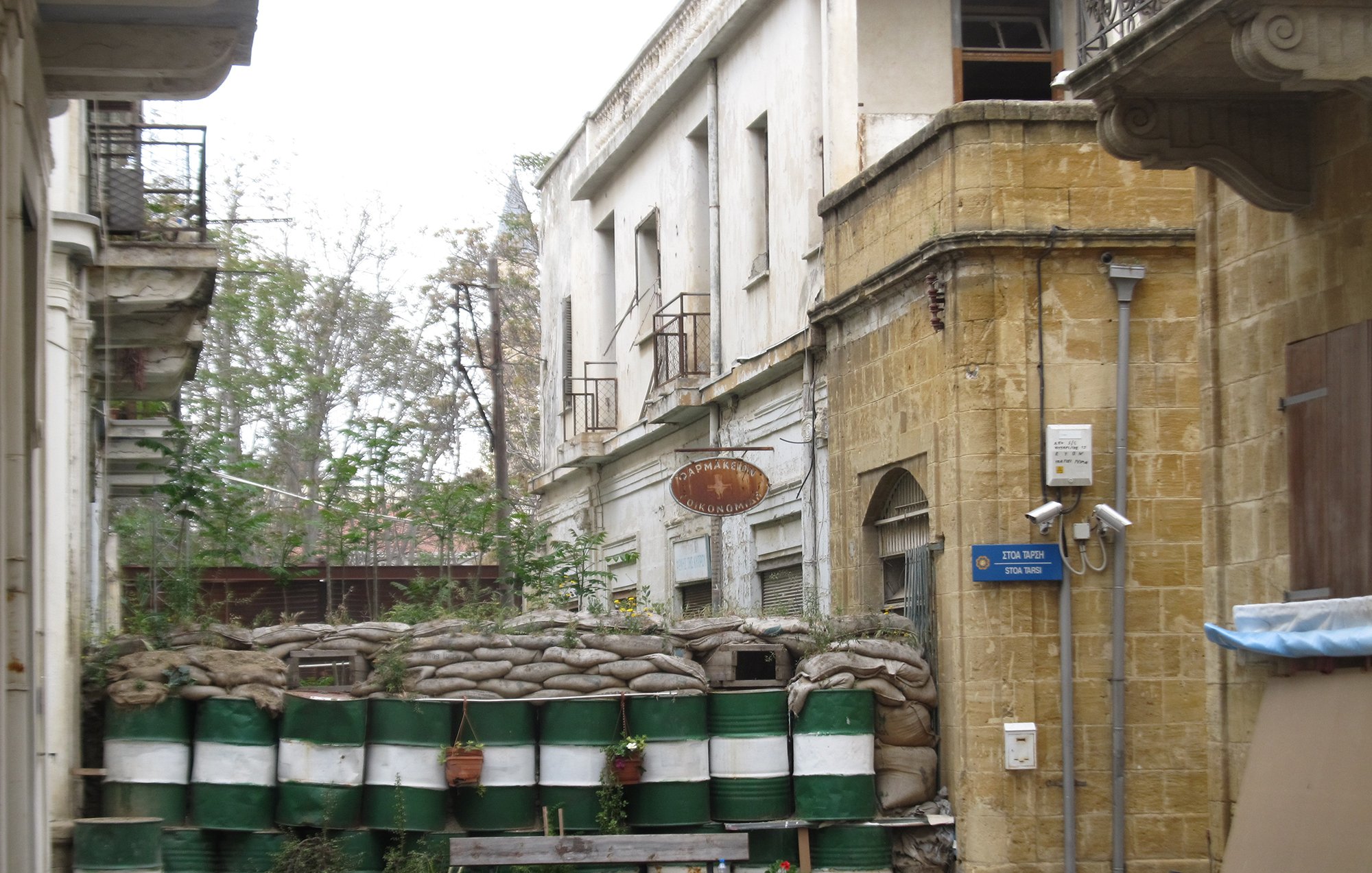 الصورة تظهر شارعاً ضيقاً في مدينة قديمة، حيث توجد مباني قديمة ذات تصميم معماري تقليدي. في نهاية الشارع، هناك حاجز مصنوع من أكياس الرمل وبراميل، مما يشير إلى وجود خطط للحماية أو الأمن. بعض النباتات تنمو حول الحاجز، مما يضفي لمسة من الحياة على المشهد. الألوان الرئيسية في الصورة هي درجات من البيج والأخضر، مما يعكس طابعاً قديماً وحالياً للمكان.