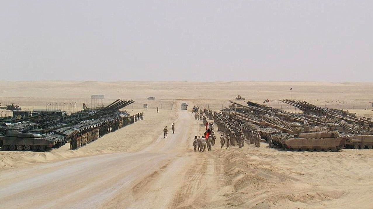 تظهر في الصورة مشهدًا صحراويًا يضم العديد من الدبابات والمركبات العسكرية. يقف عدد من الجنود في المنتصف، بينما تتواجد مركبات أخرى مصفوفة على جانبي الطريق. الخلفية تتكون من رمال الصحراء وأفق بعيد، مما يبرز الطبيعة القاحلة للمكان. تخلق الصورة انطباعًا بالقوة العسكرية والتنظيم.