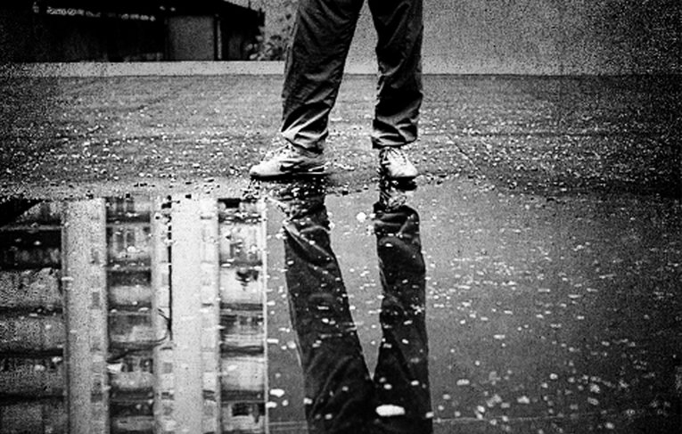 تظهر في هذه الصورة مجموعة من العناصر المثيرة للاهتمام. في المقدمة، نقف أمام شخص يرتدي بنطالًا و حذاء، واقفًا فوق سطح مغطى بالماء المتجمع، مما يشكل بركة صغيرة. يظهر انعكاس الشخص في الماء، مما يضيف بعدًا جماليًا للصورة. الخلفية عبارة عن جدار رمادي وسطح فارغ، مما يساعد على تركيز الانتباه على الشخص والانعكاس في الماء. الجو العام يبدو هادئًا ومظلمًا، مع تباين قوي بين الألوان.