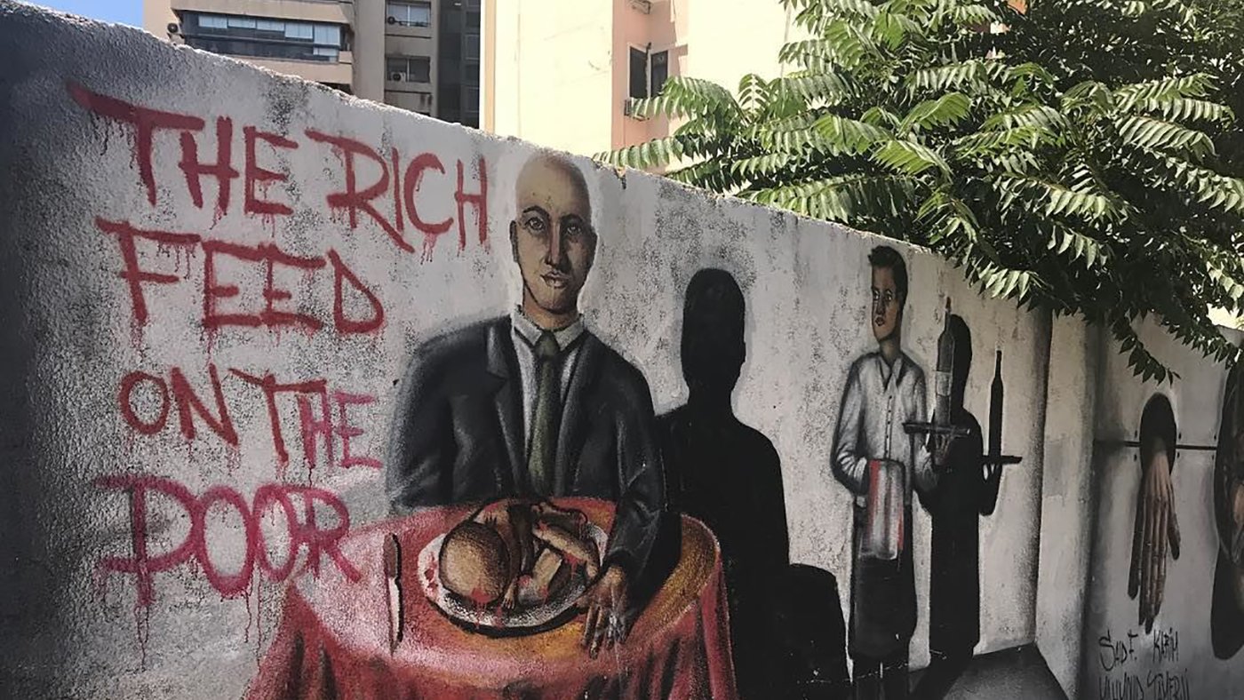 تظهر في الصورة جداريات على حائط، حيث يبرز جزء من العمل الفني رسما لشخص يجلس على طاولة، مرتديًا بدلة، ويبدو أنه يتناول الطعام. خلفه توجد ظلال لشخصيات أخرى، بما في ذلك شخص يرتدي ملابس عامل، مما يعكس تباينًا بين الطبقات الاجتماعية. تزين الجدارية أيضًا عبارة باللغة الإنجليزية "THE RICH FEED ON THE POOR" (الأغنياء يتغذون على الفقراء)، مما يوحي برسالة اجتماعية قوية حول الفوارق الطبقية والاستغلال. في الخلفية، يمكن رؤية أشجار وبعض المباني السكنية، مما يشير إلى أن هذه الجدارية تقع في منطقة حضرية.