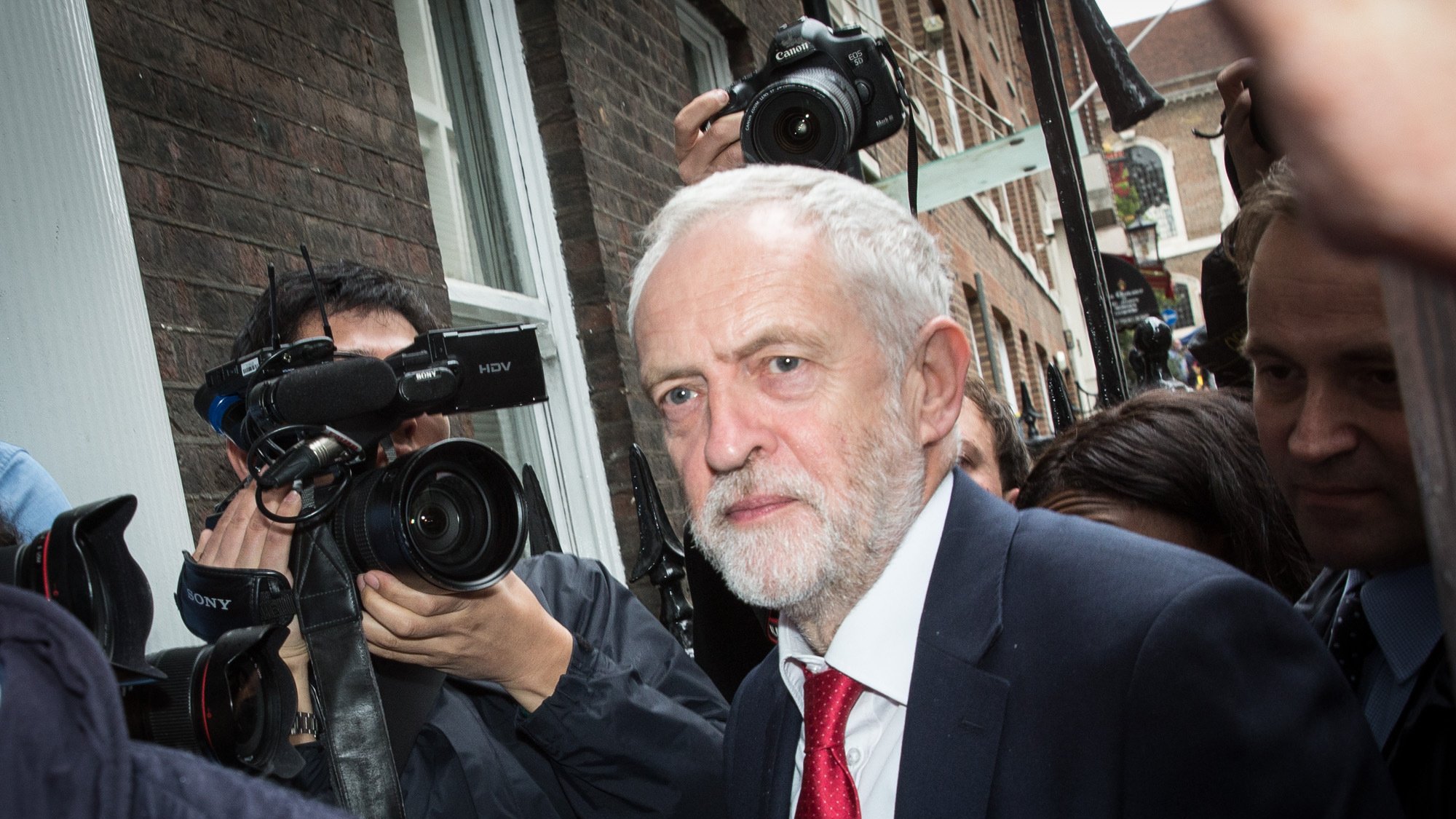 الصورة تظهر رجلًا متوسط العمر ذو لحية وشعر رمادي، يرتدي بدلة داكنة وربطة عنق حمراء. يبدو أنه في موقف يتطلب الانتباه، حيث تحيط به مجموعة من الكاميرات والصحفيين الذين يلتقطون صورًا له. الخلفية تعكس بيئة حضرية مع مبانٍ طوبية، وتعكس الحركة والاهتمام بالشخص.