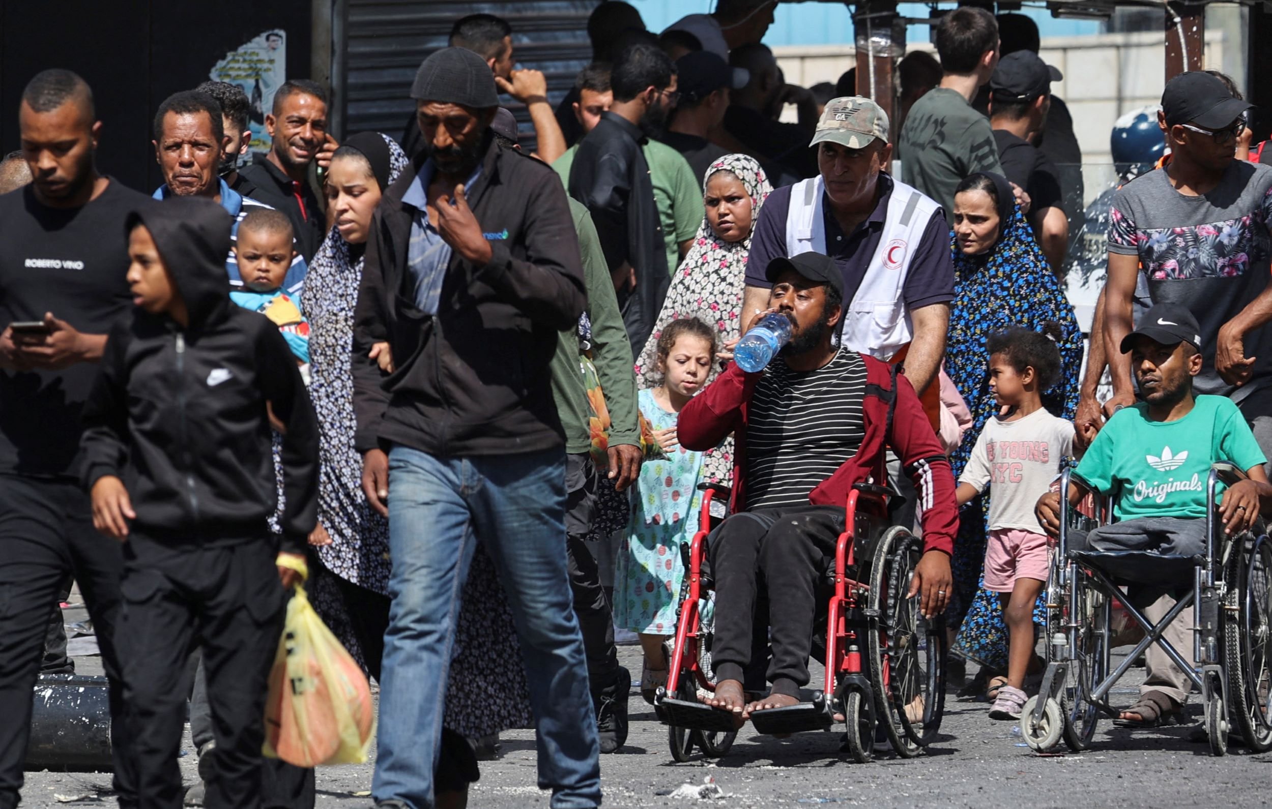 تظهر الصورة مجموعة من الأشخاص يسيرون في منطقة مزدحمة. يوجد رجال ونساء وأطفال، وبعضهم يظهر في كراسي متحركة. يتواجد في الخلفية بعض المحلات والبائعين. يبدون مشغولين أو في طريقهم إلى مكان ما، مما يعكس حركة الحياة اليومية في هذا المكان.