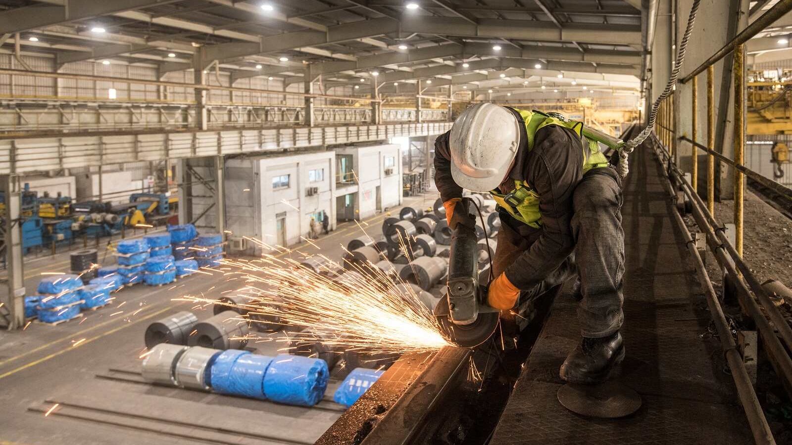 تظهر في الصورة عاملاً يرتدي خوذة وسماعات أذن، بينما يقوم بعملية قطع أو طحن المعادن في مصنع. تتطاير الشرر أثناء عملية العمل، مما يضيف إلى الحركة والديناميكية في المكان. خلف العامل، يُمكن رؤية مساحة واسعة من المصنع تضم معدات وعجلات مغطاة بأقمشة زرقاء، مما يشير إلى بيئة صناعية نشطة. الإضاءة في الصورة جيدة، مما يبرز تفاصيل العمل والبيئة المحيطة.