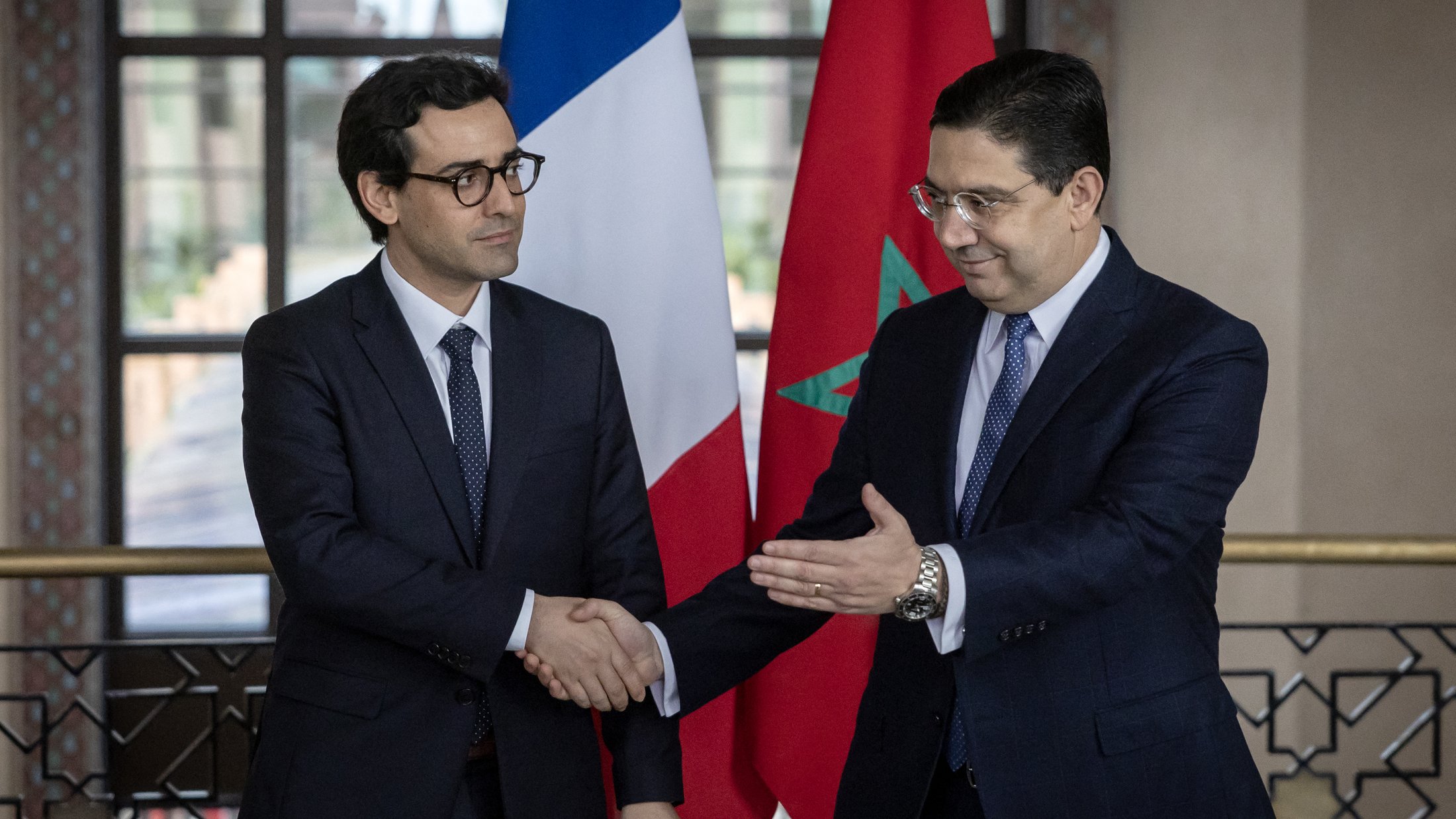 تظهر الصورة رجلين يرتديان بدلات رسمية. أحدهما يقف على اليمين ويمثل المغرب، حيث يظهر علم المغرب خلفه. الآخر يقف على اليسار ويمثل فرنسا، مع العلم الفرنسي خلفه. يبدو أن الرجلين يتصافحان في سياق رسمي، مما يشير إلى اجتماع أو تعاون بين البلدين. القاعة التي يتواجدان بها تبدو أنيقة وتحتوي على نوافذ كبيرة تسمح بدخول الضوء الطبيعي.