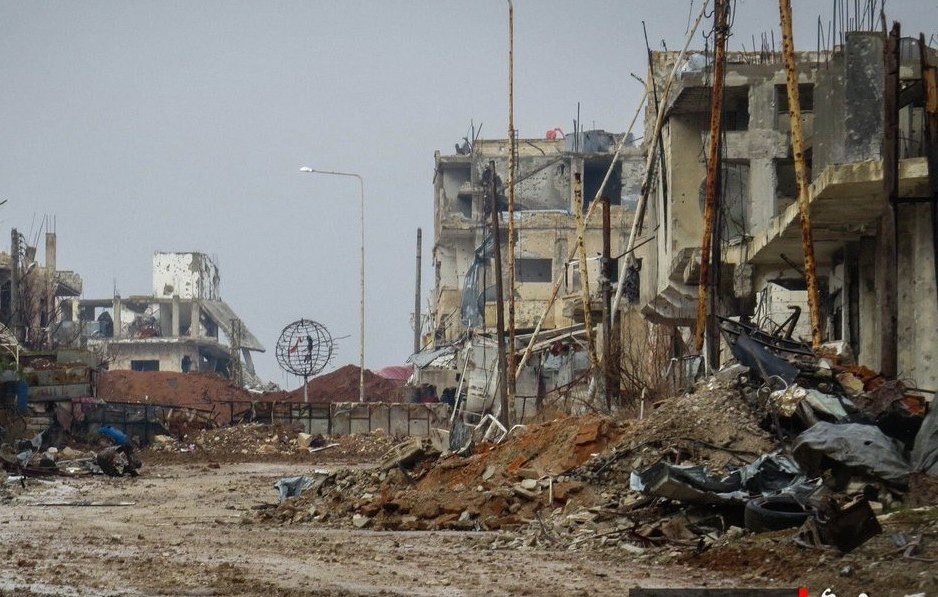 الصورة تظهر مشهدًا لمدينة مدمرة، حيث تبدو المباني متضررة بشدة، مع بقايا الهياكل المعدنية وأجزاء من الأنقاض. الطريق في المنتصف مليء بالحصى والأحجار، ويبدو غارقًا في الخراب. يظهر في الخلفية بعض الأعمدة الكهربائية التي لم تعد تعمل، مما يضيف إلى الجو القاتم والموحش للمكان. الأجواء عامة غائمة، مما يزيد من شعور الخواء واليدانة في هذا المشهد.