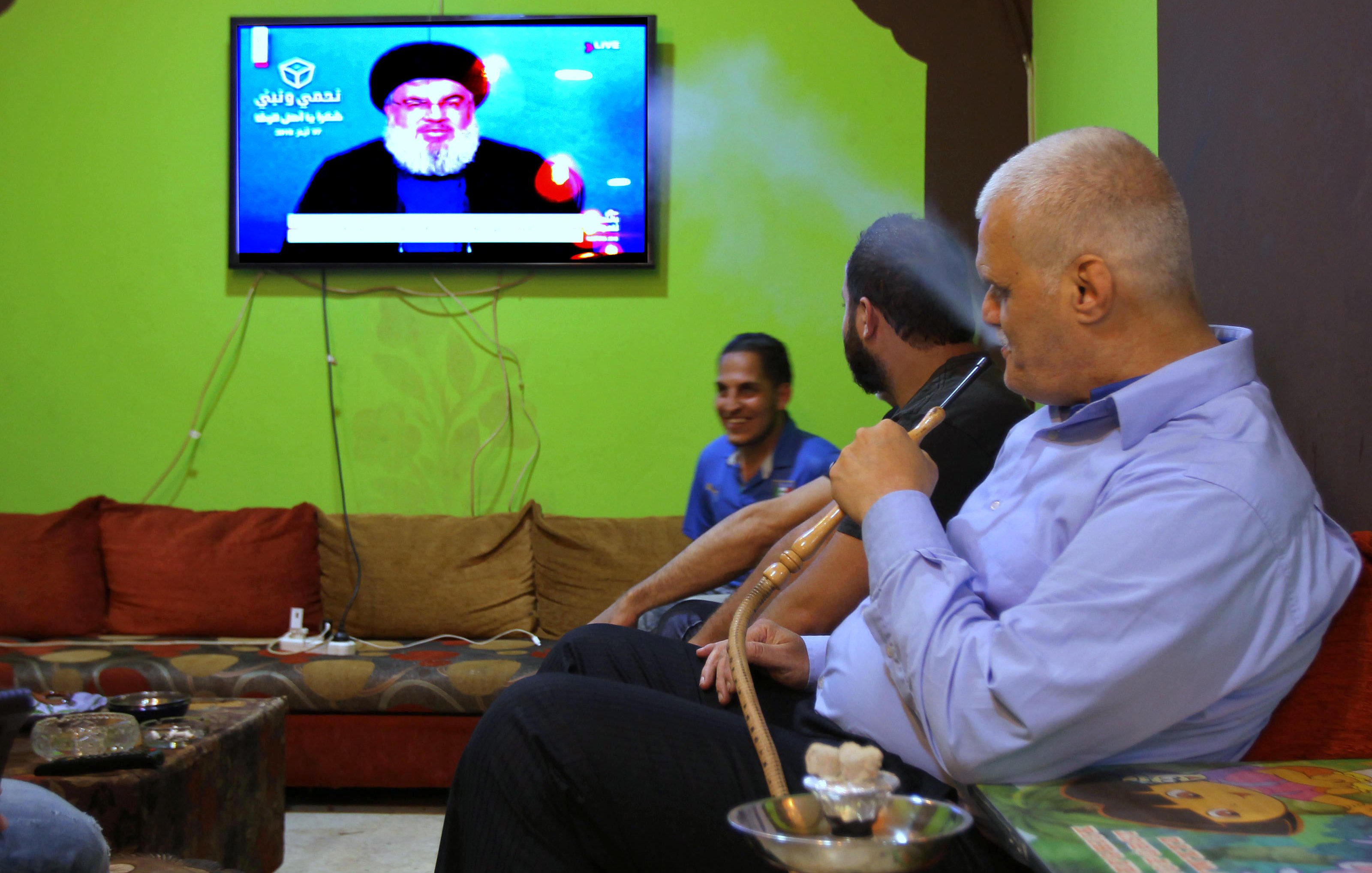 تظهر في الصورة مجموعة من الأشخاص يجلسون في غرفة معيشة مريحة، حيث يجلس رجلٌ على الأريكة وهو يدخن الشيشة. خلفه، يظهر تلفاز يُبث عليه صورة لشخص معروف يتحدث. الجدران تؤدي إلى أجواء دافئة، بينما الركن الآخر يُظهر شخصًا ضاحكًا. الألوان في المكان تتنوع بين الأخضر والبني، مما يعطي شعورًا بالراحة.