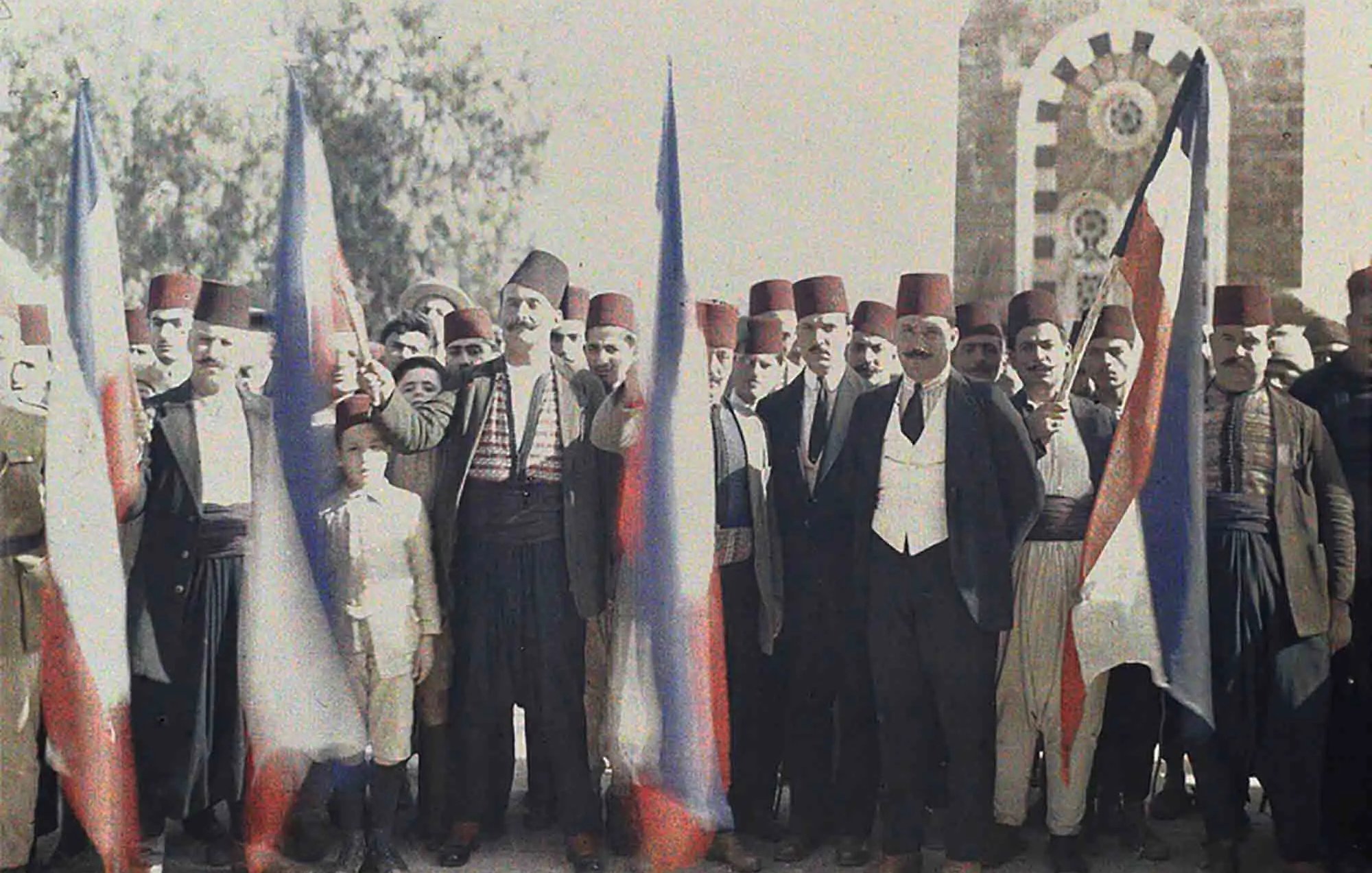 تظهر في الصورة مجموعة من الرجال واقفين في صف واحد، يحملون أعلامًا. يرتدي معظمهم الملابس التقليدية، بما في ذلك الأزياء التي تضم الطربوش الفاسي. يظهر في الصورة أيضًا صبي صغير في وسط المجموعة. يبدو أنهم يحتفلون أو يشاركون في فعالية معينة، حيث يُظهر تعبيرهم الحماس والجدية. الخلفية تحتوي على مبنى قديم، مما يعطي انطباعًا تاريخيًا ومناسبًا للسياق.