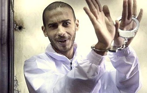 تظهر في الصورة شخصًا يرتدي زيًا أبيض يبدو كأنه في أحد أماكن الاحتجاز. يده مقيّدة بالأصفاد، ويبدو أنه يعبر عن فكرة أو شعور ما من خلال تعابير وجهه وحركات يده. الإضاءة والبيئة المحيطة تشير إلى مكان مغلق، مما يعزز الإحساس بالاحتجاز.