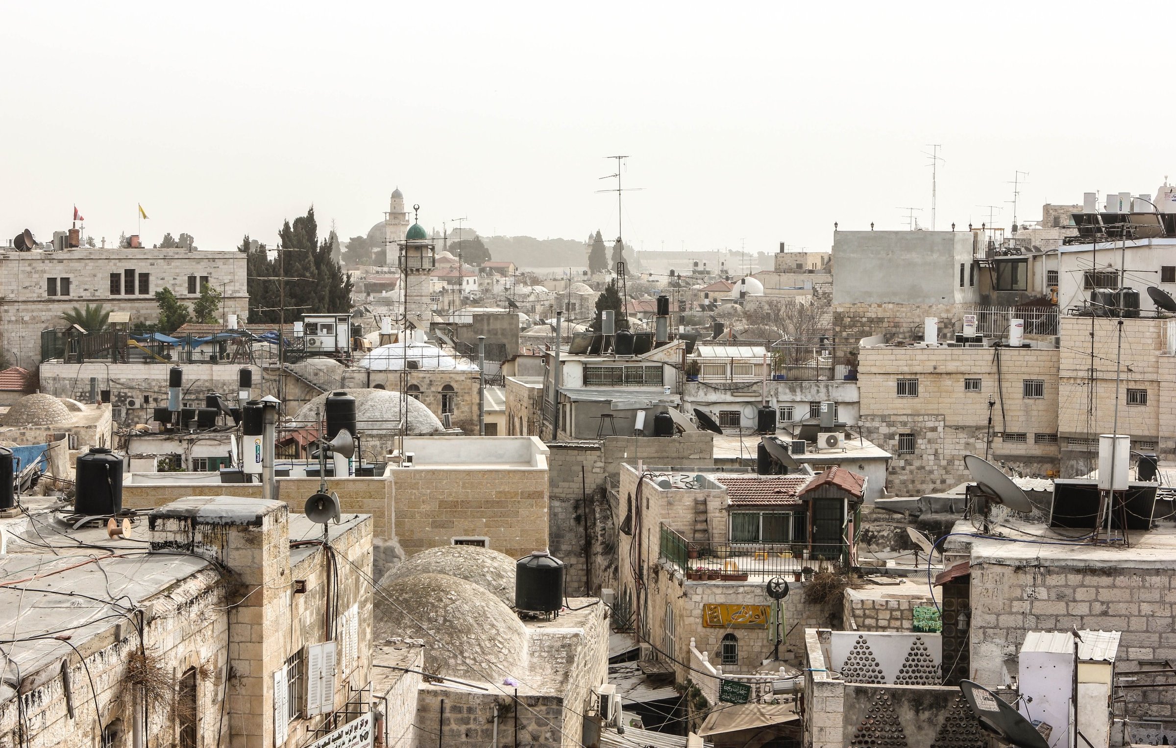 الصورة تُظهر منظرًا لأحد الأحياء القديمة، حيث تتداخل المباني التقليدية مع الأسطح المسطحة وأجهزة التكييف. يوجد أيضًا بعض الأشجار ومآذن المساجد في الخلفية، مما يشير إلى طابع تاريخي وديني للمنطقة. السماء تبدو مائلة إلى الرمادي، مما قد يدل على أجواء غائمة أو ضبابية.