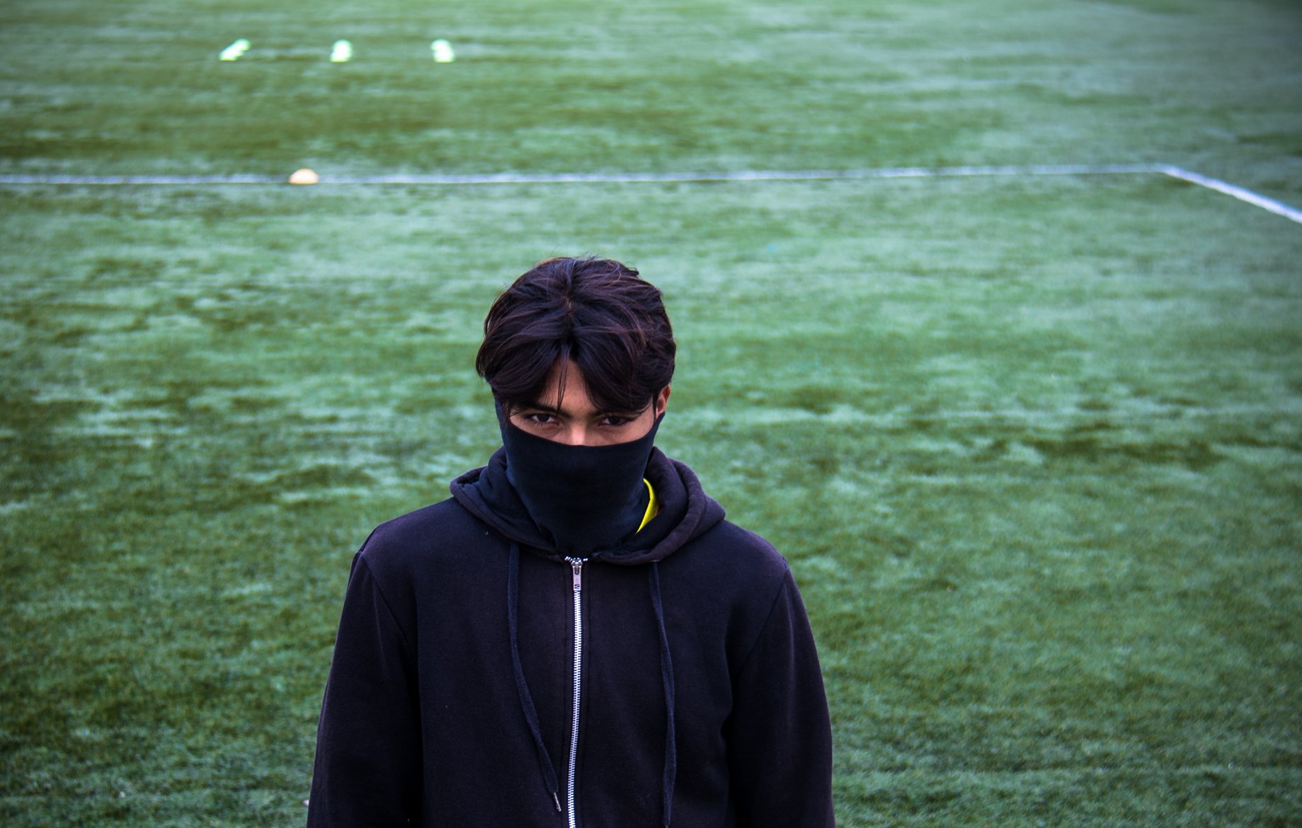 تظهر في الصورة شخصية شاب يقف على أرضية ملعب رياضي مغطاة بالعشب الصناعي. يرتدي الشاب سترة داكنة، ويغطي وجهه بوشاح أو قطعة قماش سوداء، مما يضفي عليه مظهرًا غامضًا. ينظر مباشرة إلى الكاميرا، مما يبرز قوة تعبيره. الخلفية تتكون من اللون الأخضر للأرضية الرياضية، مع بعض العلامات أو الأعلام الموزعة عليها.