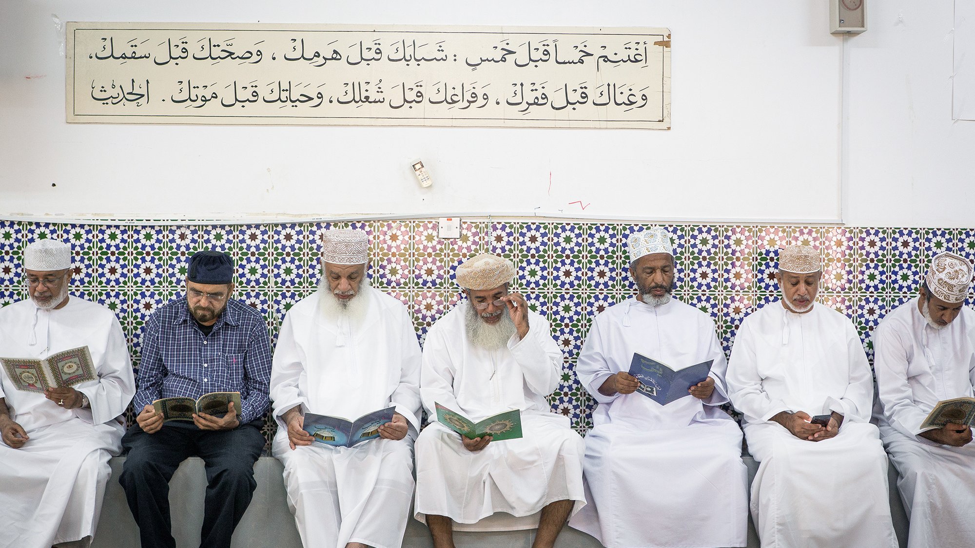 تظهر في الصورة مجموعة من الرجال يجلسون في صف واحد داخل مكان يبدوا أنه مسجد أو قاعة صلاة. كل منهم يحمل كتاباً، ويبدو أنهم يقرؤون أو يتدارسون. الجدران مزينة ببلاط ملون مزخرف، وهناك لوحة نصية مثبتة في الأعلى تحتوي على آيات أو نصوص دينية. يظهر في الصورة أجواء من التركيز والسكينة، مع تعبيرات الهدوء على وجوههم.