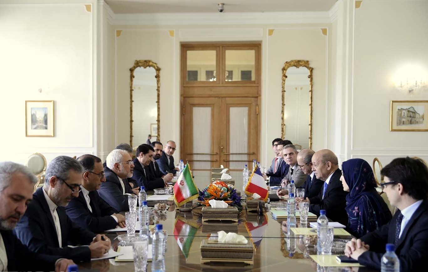 تظهر في الصورة قاعة اجتماعات تجمع وفدين من رجال السياسة. يجلس الأعضاء على طاولة طويلة مرتبة بشكل منظم، حيث توجد أعلام إيران وفرنسا على الطاولة. يتواجد في الجانبين رجال ونساء يرتدون ملابس رسمية، ويبدو أن النقاشات جادة ومتبادلة. تعكس الأجواء في الغرفة رسمية واحترافية، مع تفاصيل معمارية متقنة في المكان.