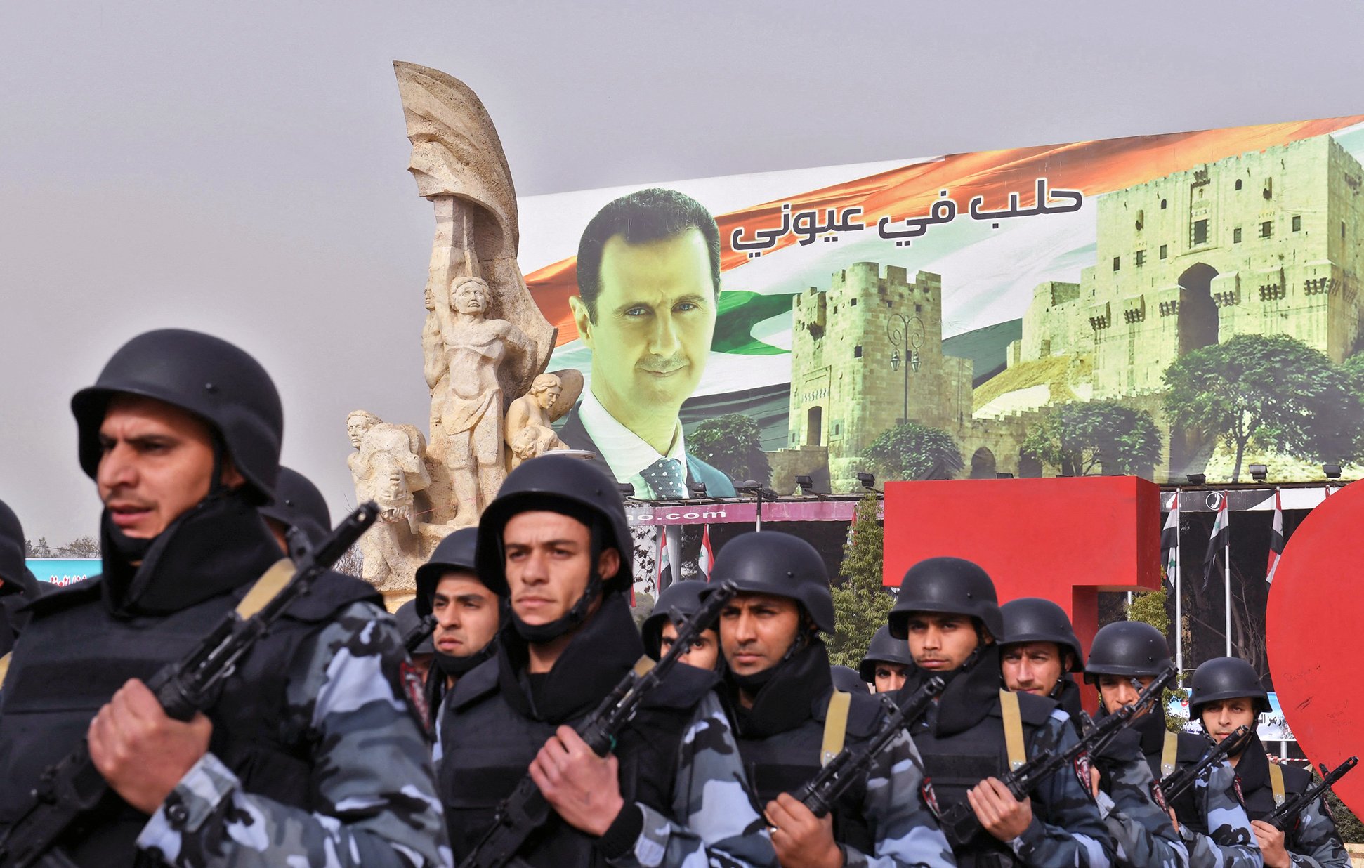 تظهر الصورة عرضًا عسكريًا أو تظاهرة، حيث يسير مجموعة من الجنود في صفوف منظمة، يرتدون زيًا عسكريًا ويحمون أسلحة. خلفهم يوجد لافتة ضخمة تحمل صورة لرجل، تترافق مع مشاهد معمارية تتعلق بسوريا. تظهر أيضًا بعض الرموز الوطنية مثل الأعلام. الأجواء تعكس نوعًا من القوة والتأييد السياسي.