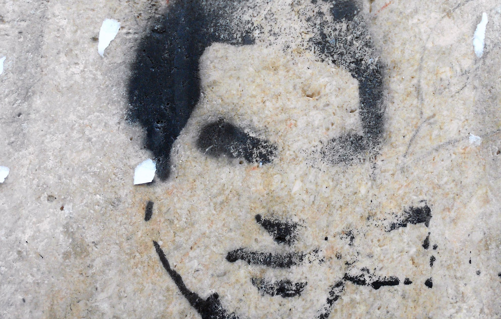 الصورة تظهر رسومًا جدارية تُظهر وجه إنسان بأسلوب الطلاء الرش. يبدو أن الرسم مقطوع بعض الشيء، مع تفاصيل غامضة لفم وأنف وعينين. الخلفية غير منتظمة وقد تشتمل على بعض آثار الندوب أو التآكل، مما يعطي الانطباع بأنها قد رسمت على سطح قديم أو متآكل. اللون المستخدم في الرسمة أسود بينما الخلفية ذات لون فاتح.