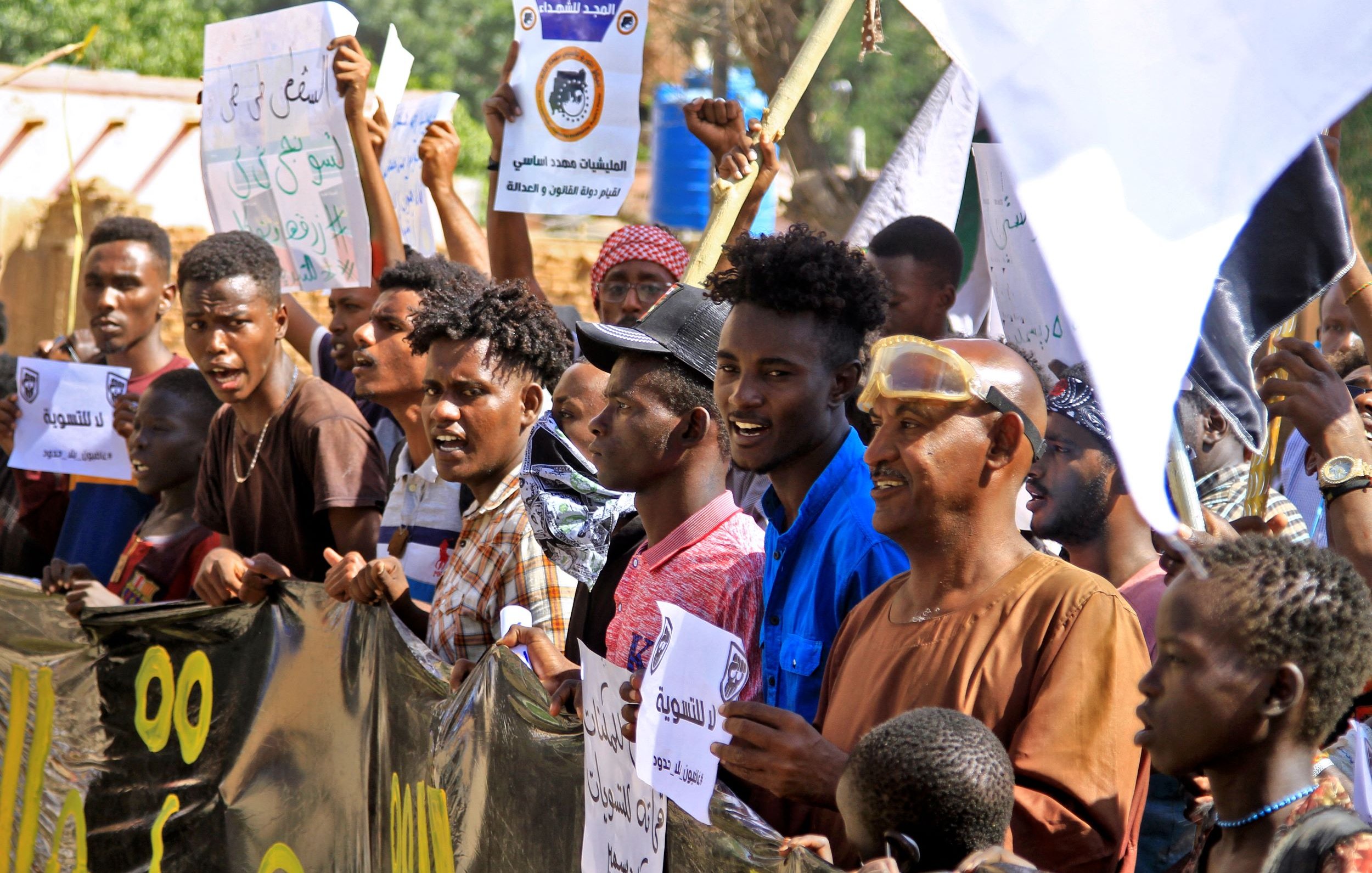 الصورة تظهر مجموعة من الناس في تظاهرة، يحملون لافتات و شعارات. يبدو أنهم متحمسون ويعبرون عن آرائهم. يظهر في الخلفية بعض المباني والأشجار، مما يدل على أن التجمع يحدث في منطقة مفتوحة. الوجوه تعكس مشاعر مختلفة، مثل الحماس والعزيمة.