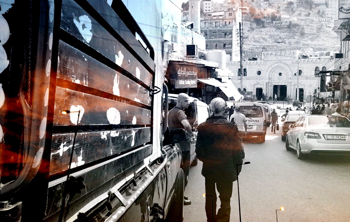 تبدو الصورة عبارة عن مشهد حضري يظهر شارعًا مزدحمًا حيث يسير أحد الأشخاص بعصا مساعدة. تظهر في الخلفية مباني متعددة الأشكال، وبعض السيارات تسير في الشارع. الأجواء تعكس حركة الحياة اليومية، ويبدو اللون الرمادي والأسود مسيطرًا مع لمسات من الألوان الدافئة، مما يضفي شعورًا بالحنين أو الذكريات.