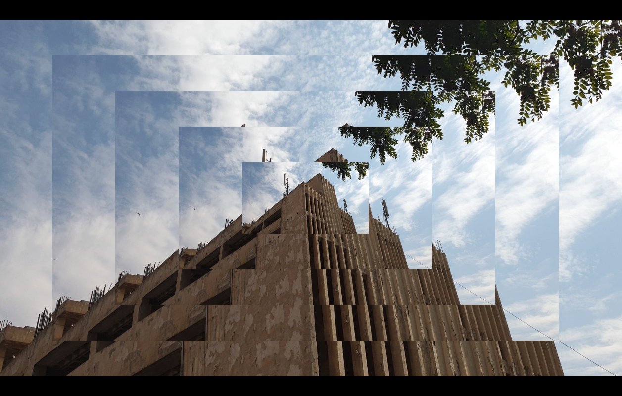 تظهر الصورة مشهداً لمبنى معاصر يظهر فيه تصميم معماري مميز، حيث يتكون المبنى من عدة طبقات وأشكال هندسية واضحة. السماء خلف المبنى محاطة بسحب بيضاء متناثرة، مما يضيف جمالاً إلى المشهد. في الزاوية العليا، هناك مجموعة من الأوراق الخضراء لشجرة تضفي لمسة طبيعية على الصورة. التصميم يبدو مزيجاً من الحداثة والطبيعة، مما يخلق تبايناً جميلاً بين العمارة والبيئة المحيطة.