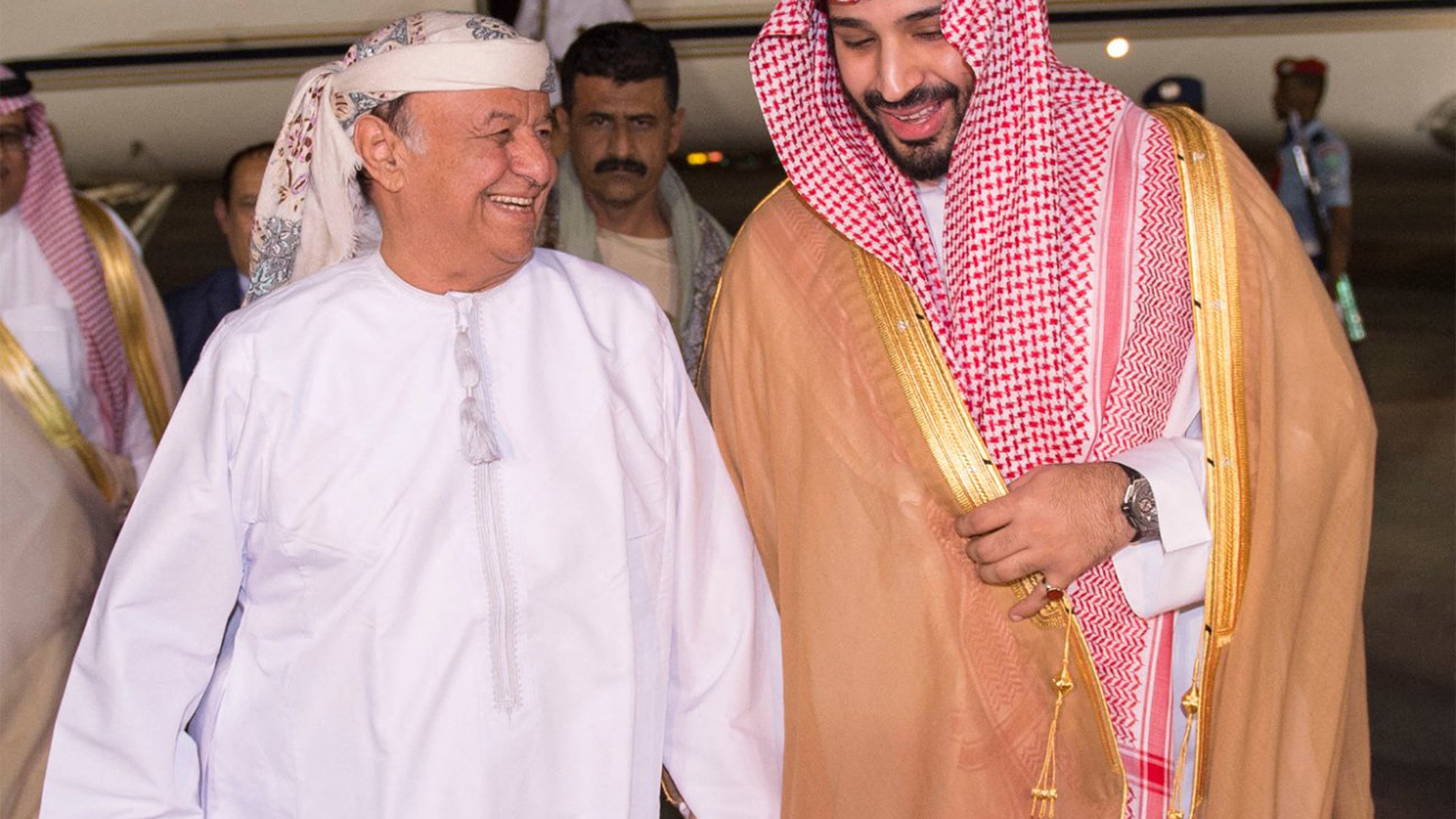 في الصورة، يمكن رؤية رجلين يتحدثان معاً بمرح. يرتدي أحدهما زياء تقليدية عربية مكونة من جبة بيضاء وعقال، بينما يرتدي الآخر الملابس السعودية التقليدية، تشمل العباءة والكوفية. يظهر خلفهم طائرة خاصة، مما يشير إلى وصولهم إلى مكان محدد. تبدو الأجواء مفعمة بالترحاب والود.