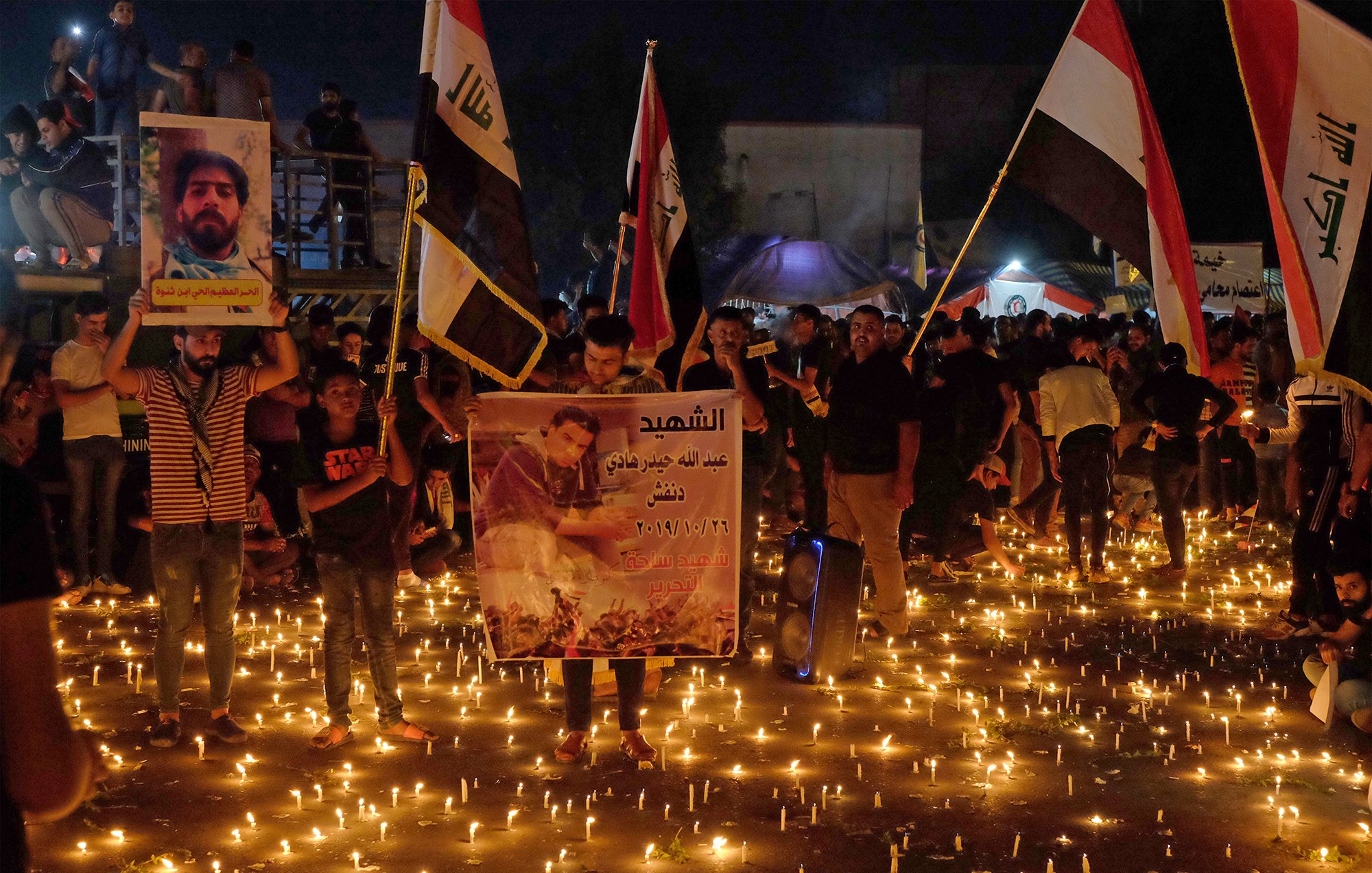 توضح الصورة تجمعًا حاشدًا للناس في مكان مفتوح خلال الليل، حيث يضيء المكان بكثرة الشموع الموزعة على الأرض. يحمل المشاركون لافتات وصورًا، وبعضهم يرفع أعلامًا تمثل بلدانهم. الجو يبدو مهيبًا، حيث يعبر الناس عن حزنهم أو إحياء ذكرى معينة، مما يعكس شعورًا بالوحدة والتضامن في هذه اللحظة.