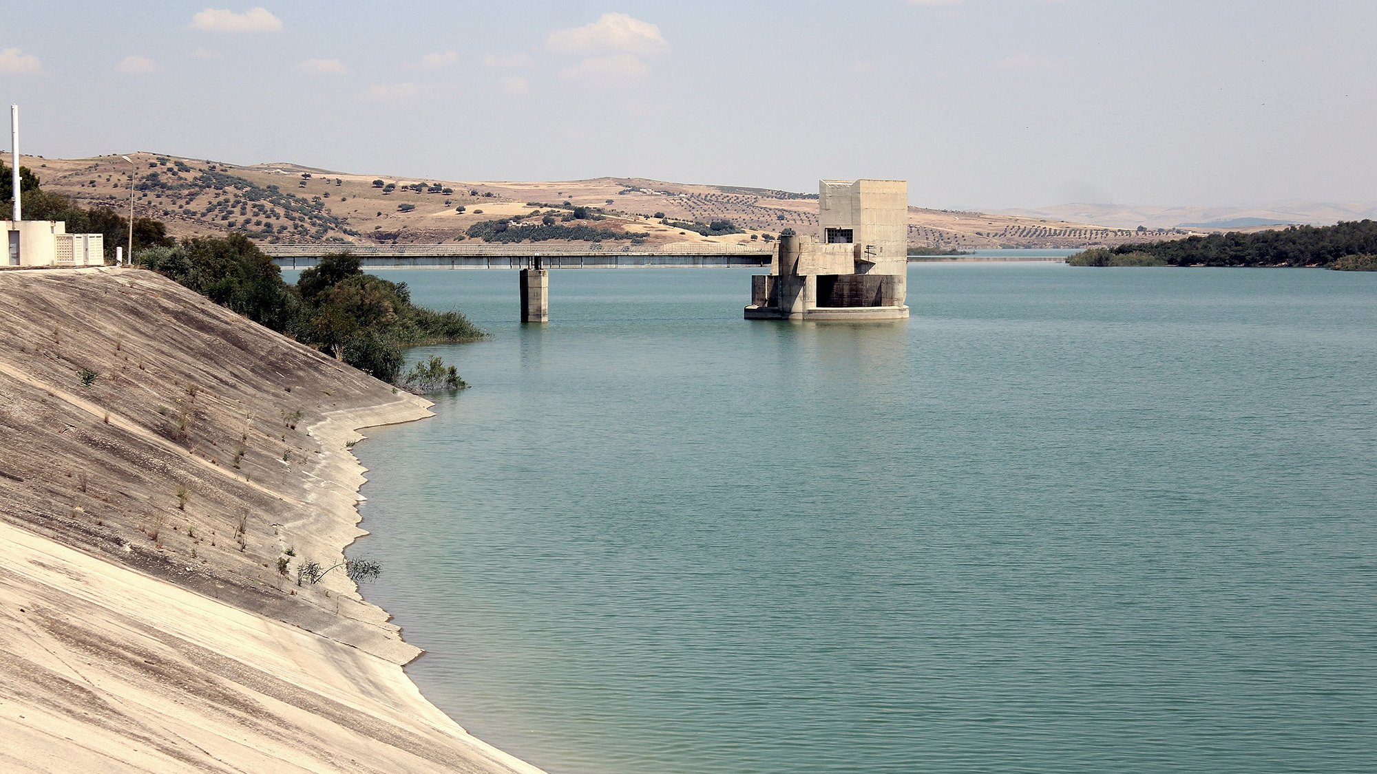 تظهر الصورة منظرًا هادئًا لبحيرة محاطة بتضاريس جبلية. يتوسط المشهد بناء هندسي يُحتمل أن يكون محطة لتحكم في المياه أو وسيلة لتخزينها. المياه باللون الأزرق الفاتح تعكس السماء، بينما تمتد ضفاف البحيرة بلطف، مع وجود تدرجات من اللون الرمادي الفاتح في الرمال. الأفق يكتمل بكتل من التلال الخضراء البعيدة، مما يضفي على الصورة جمالًا طبيعيًا وهدوءً.