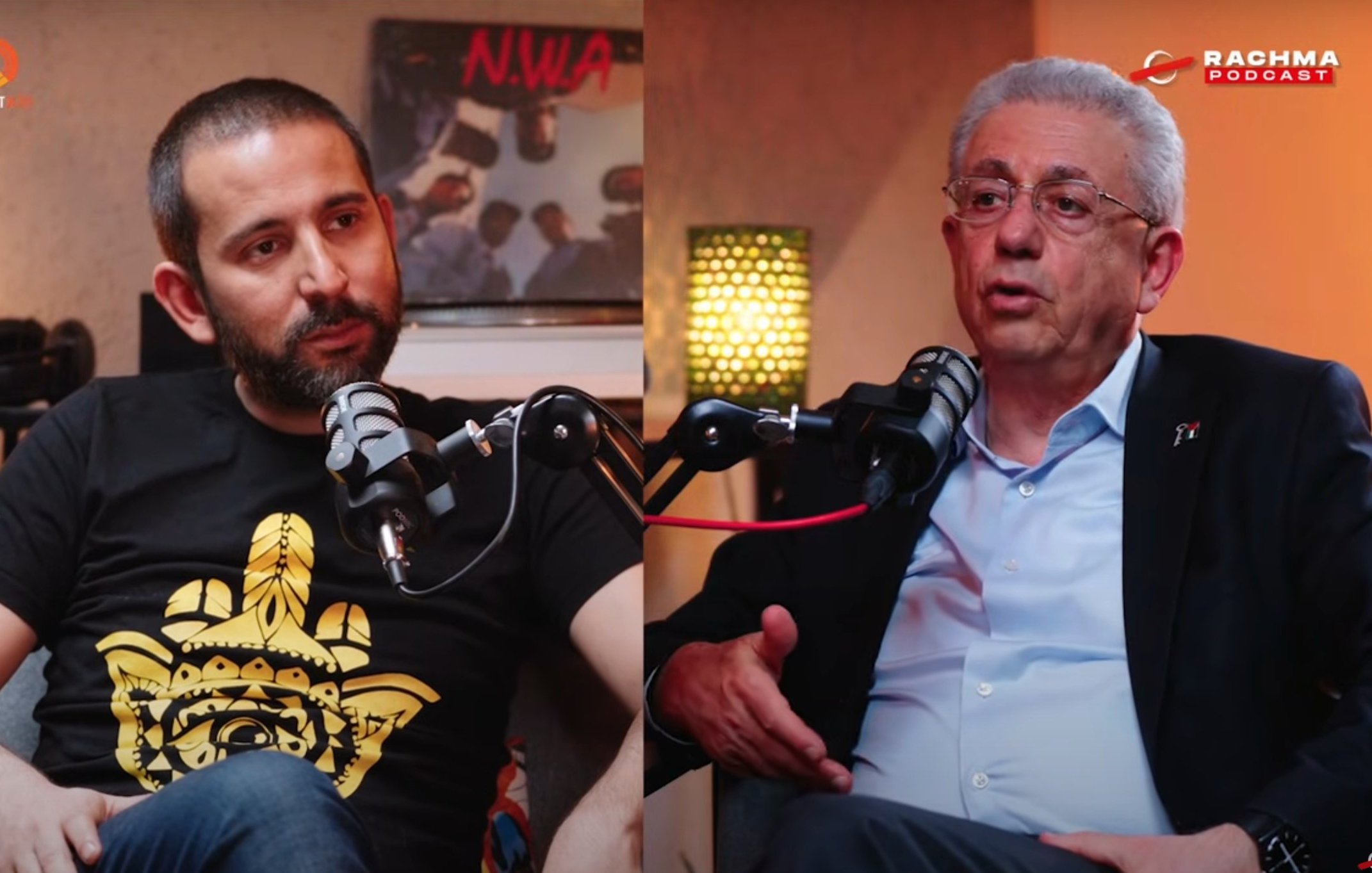 في الصورة، يظهر شخصان يجلسان معاً، يتحدثان، وأمام كل منهما ميكروفون.