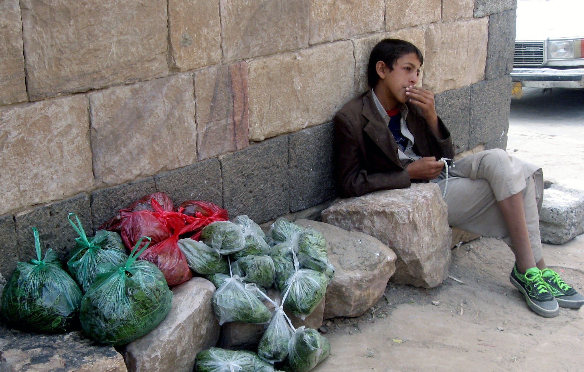 في الصورة، يظهر فتى جالساً بجوار حائط حجري. يتكئ على صخرة كبيرة وينظر إلى المسافة. يرتدي ملابس بسيطة، ويبدو أنه في حالة تفكير أو انتظار. بالقرب منه، توجد أكياس ملونة تحتوي على خضروات، بعضها بالأخضر والآخر باللون الأحمر، موضوعة بشكل منظم على الأرض. تعكس الصورة أجواء حياة الشارع وبساطة الحياة اليومية.