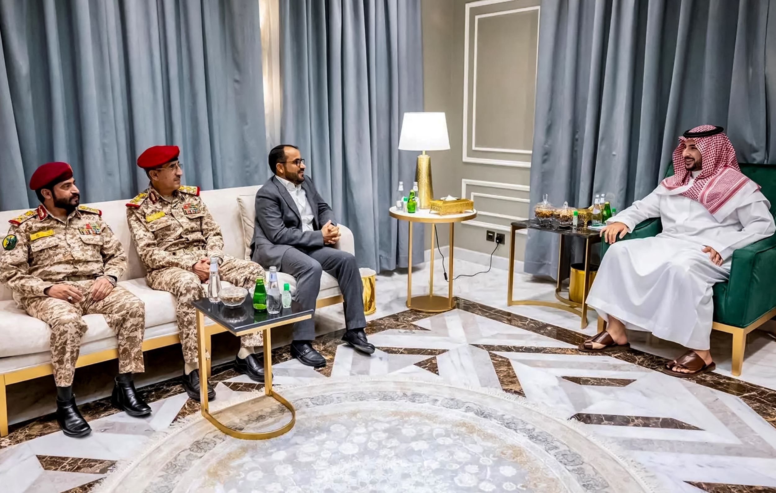 تظهر الصورة مجموعة من الأشخاص الذين يجلسون في غرفة معيشة أنيقة. في الجهة اليسرى، يوجد ضابطان عسكريان يرتديان زيًا عسكريًا، أحدهما يرتدي قبعة حمراء. بينما يجلس رجل ثالث في المنتصف بزي مدني. على اليمين، يوجد رجل يرتدي لباسًا تقليديًا سعوديًا (ثوب وغطاء رأس) يجلس على أريكة. يوجد أيضًا طاولة صغيرة أمامهم تحتوي على بعض المشروبات. الجدران مكسوة بستائر تتناسب مع الديكور الداخلي.