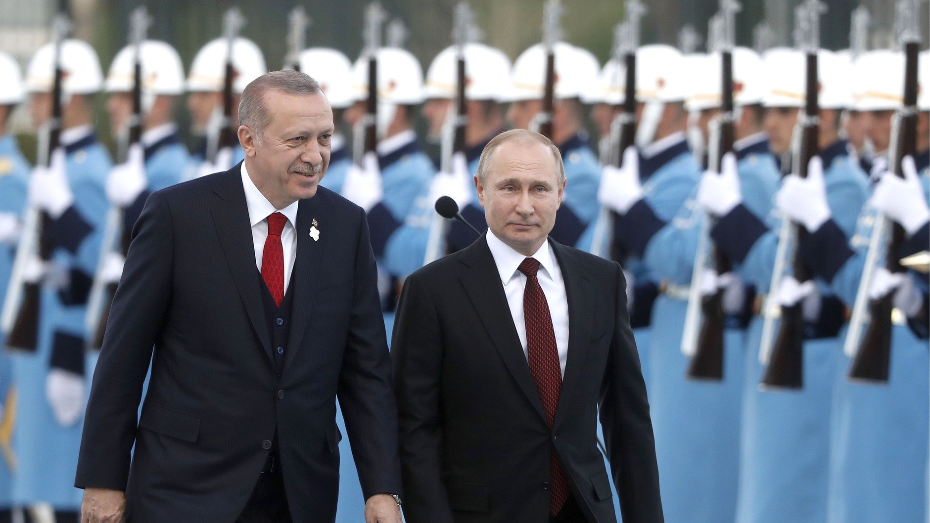 الصورة تظهر شخصين يسيران معًا، بينما خلفهما يظهر مجموعة من الحراس يرتدون زيًا موحدًا، ويقفون في صفوف منتظمة. الشخص الأول يرتدي بدلة داكنة مع ربطة عنق حمراء، بينما الثاني يرتدي بدلة أيضًا ولكن بلون مختلف. يبدو أن المشهد يتمثل في مراسم رسمية أو استقبال، حيث تم ترتيب الحراس بشكل أنيق. الجو العام بالصورة يوحي بالتعاون أو اللقاء بين الشخصيتين.