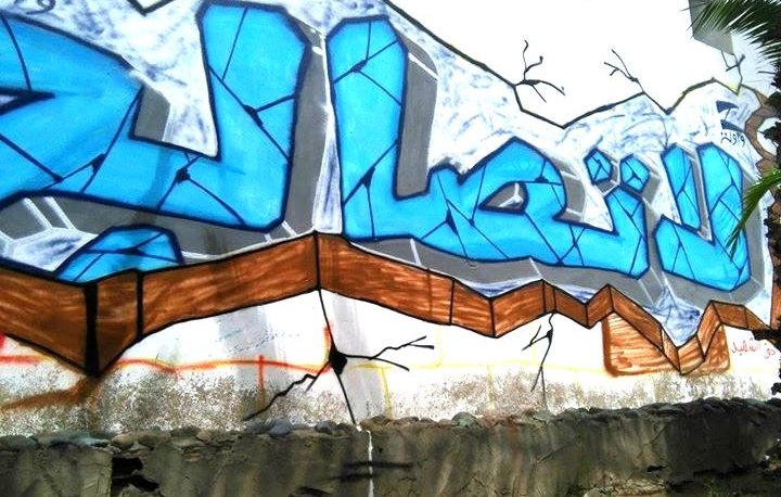الصورة تظهر جدارية من فن الجرافيتي على حائط. الكلمة المكتوبة باللون الأزرق الفاتح تتوسط الجدارية، وتحيط بها بعض الخطوط والتفاصيل باللون الأسود. يبدو أن الجدارية تجذب الانتباه بلونها الزاهي وتصميمها الفني. الحائط يظهر بعض التشققات، مما يضيف إلى الجمالية الفنية للجدارية محاطة بالعشب والأوساخ في الأسفل.