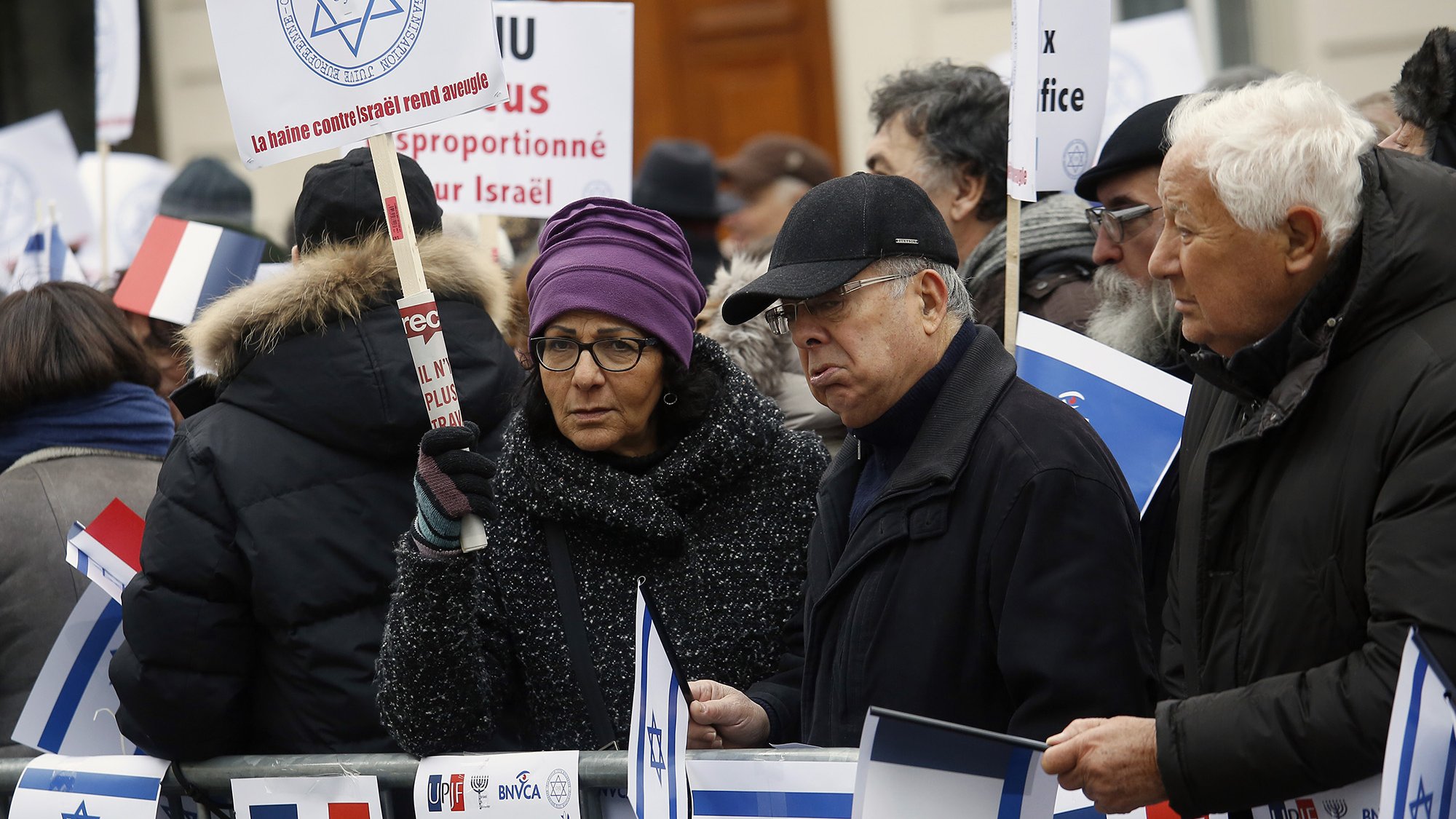 الصورة تظهر مجموعة من الأشخاص في تجمع أو مظاهرة. يحملون لافتات وأعلام، من بينها العلم الإسرائيلي، ويرتدون ملابس شتوية. يبدو أن المتظاهرين يعبرون عن آرائهم حول قضية معينة تتعلق بإسرائيل. المشهد يعكس تفاعل الناس مع قضايا سياسية واجتماعية.