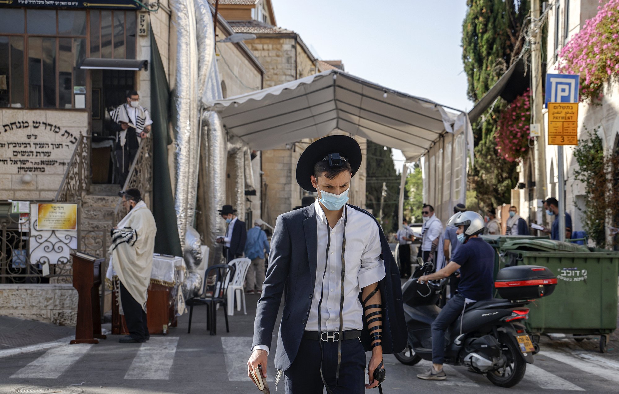 تظهر الصورة شابًا يرتدي بدلة مع قبعة، وهو يحمل هاتفًا محمولًا في يده. يتزين ذراعه بلفائف التوراة (تيفيلين)، ويبدو أنه في أحد الأماكن العامة. في الخلفية، توجد مجموعة من الأشخاص، وبعضهم يرتدي ملابس تقليدية، كما يظهر خيمة كبيرة على جانب الطريق. تتميز الصورة بأجواء حضرية مع وجود نباتات وبيئات سكنية.