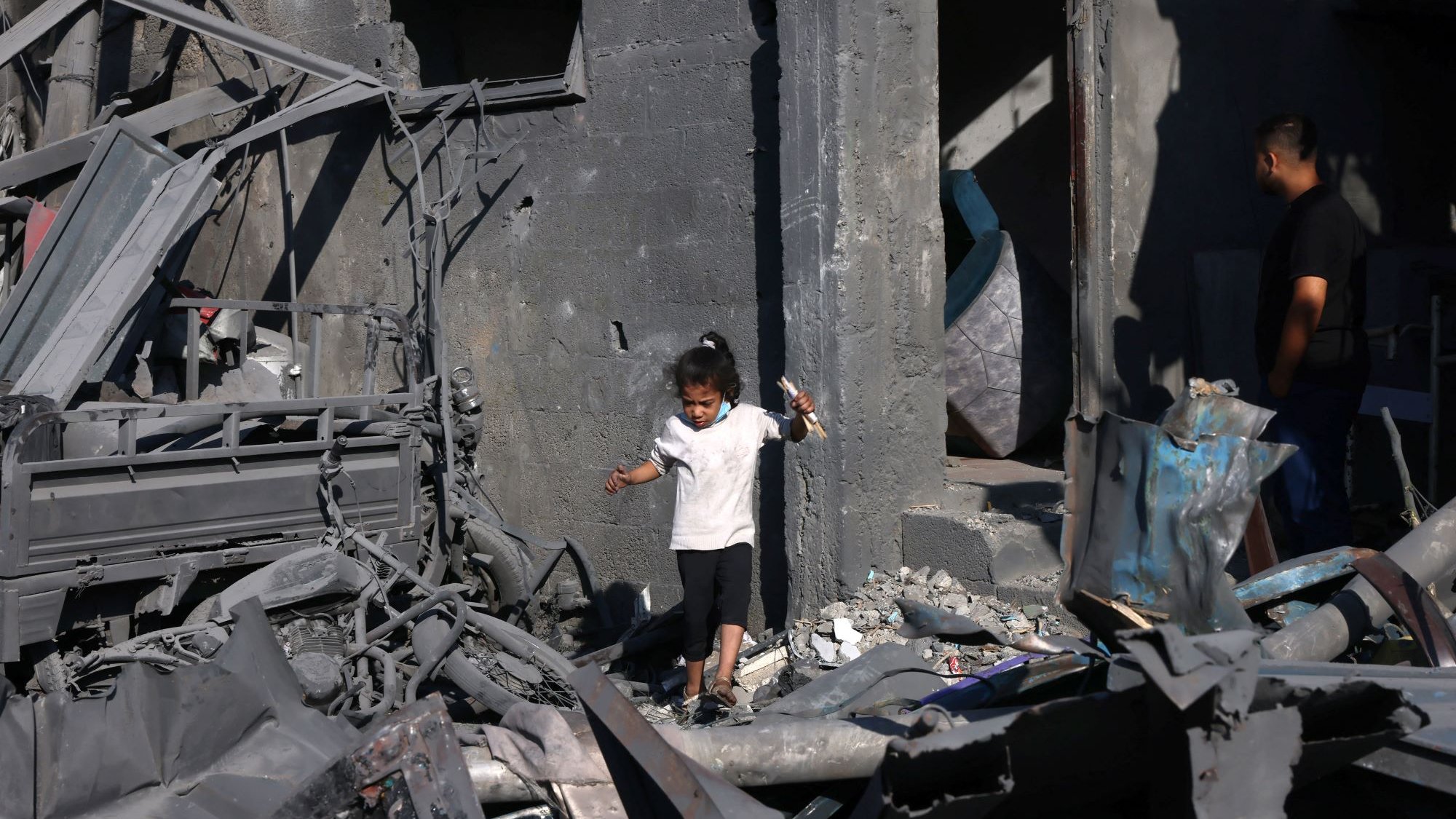 تظهر الصورة طفلاً صغيراً يرتدي الملابس البيضاء وهو يسير بين أنقاض مبنى مدمّر. الجدران حوله متهدمة ومغطاة بالرماد، مما يدل على الدمار الذي حل بالمكان. في الخلفية، يمكن رؤية شخص بالغ يقف بالقرب من الباب، يبدو وكأنه يتفحص وضع المكان. الأجواء تعكس حالة من الحزن والفوضى نتيجة الكارثة.