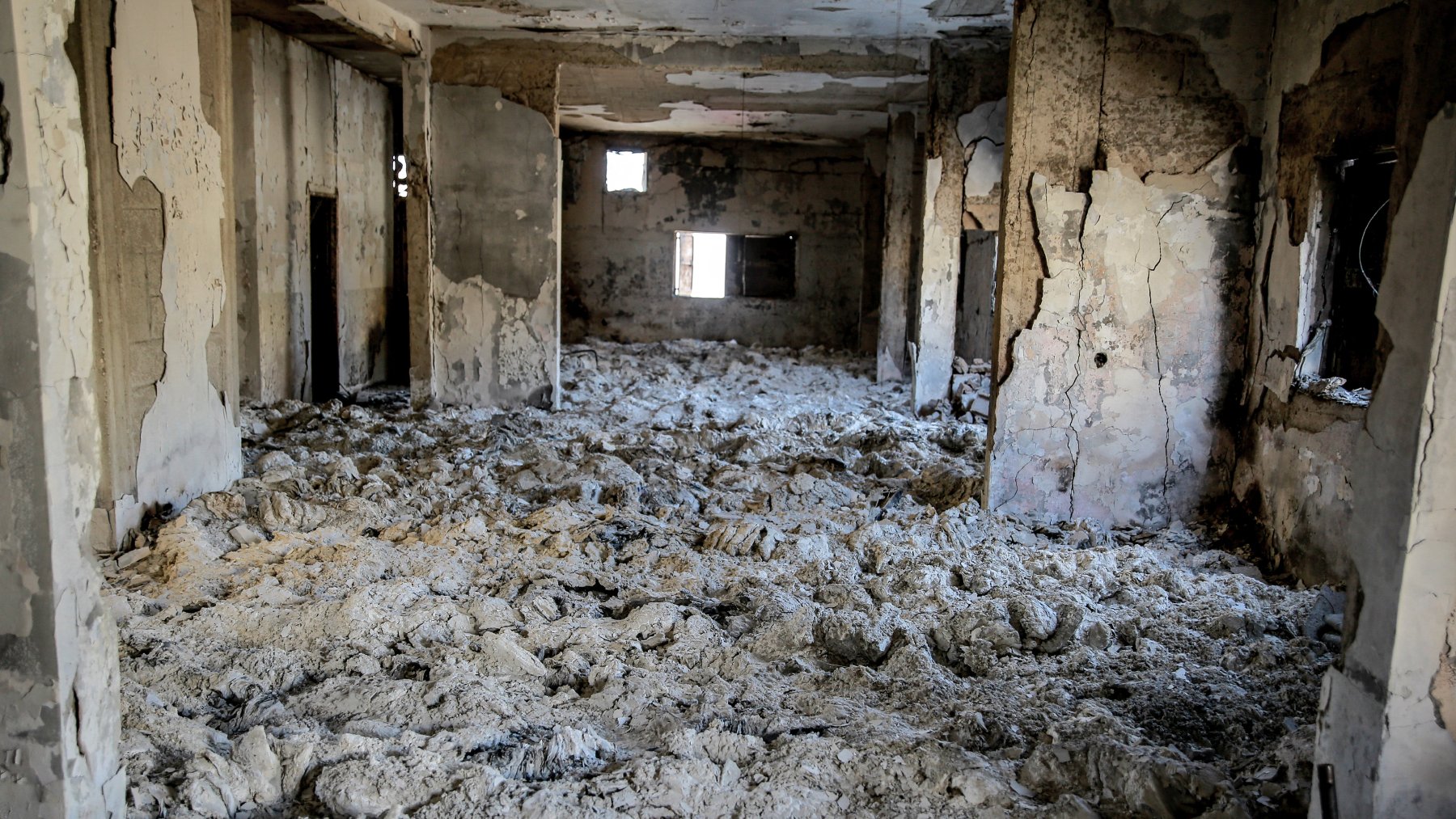 تظهر الصورة غرفة داخل مبنى تضرر بشكل كبير، حيث الجدران والسقف متهدمان، والأرضية مغطاة بطبقة من المواد البيضاء، مما يوحي بتدهور شديد في حالة المبنى. يدخل الضوء من بعض النوافذ المكسورة، مما يضيف شعورًا بالخراب والمهجورية إلى المكان.