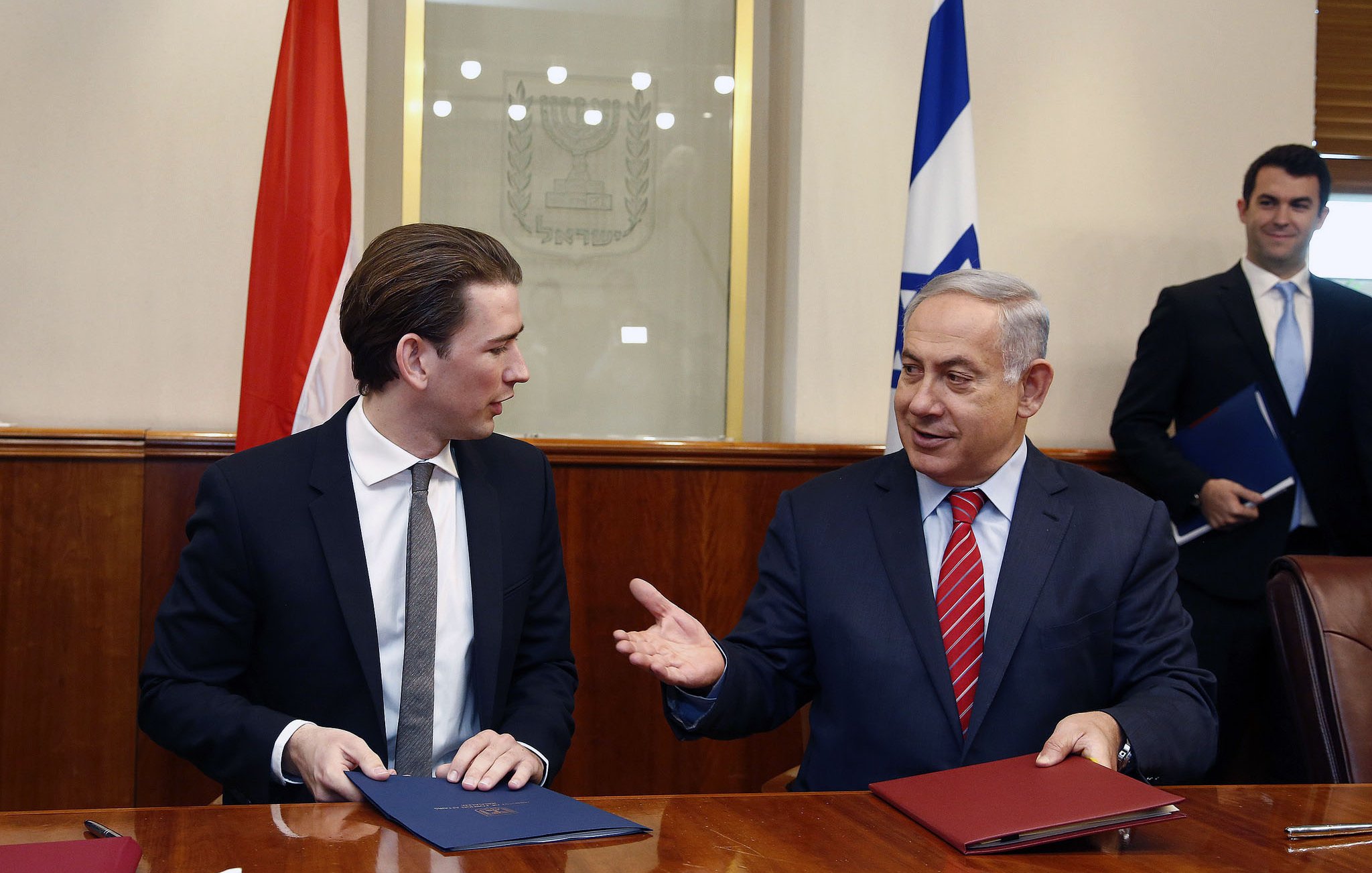 الصورة تظهر مشهداً من اجتماع رسمي، حيث يجلس شخصان على طاولة. الشخص الأول يرتدي بدلة داكنة ولديه شعر قصير، بينما الشخص الثاني يرتدي بدلة مع ربطة عنق حمراء. يبدو أن كلاهما مشارك في نقاش أو اتفاقية رسمية، حيث يوجد خلفهم علمين. الجو العام يبدو جاداً ومهنياً.