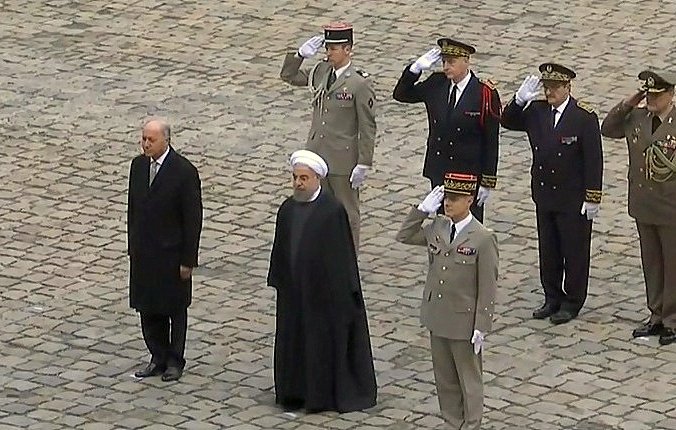 الصورة تظهر مجموعة من الأشخاص في موقف رسمي. يوجد شخصان في المقدمة، أحدهما في بدلة رسمية والآخر يرتدي الزي التقليدي. خلفهم يوجد عدد من الضباط العسكريين بزيهم الرسمي، يقومون بتحية عسكرية. الأرضية مكونة من حجارة مرصوفة. الجو يبدو رسميًا وجادًا، مما يشير إلى حدث ذو أهمية سياسية أو عسكرية.