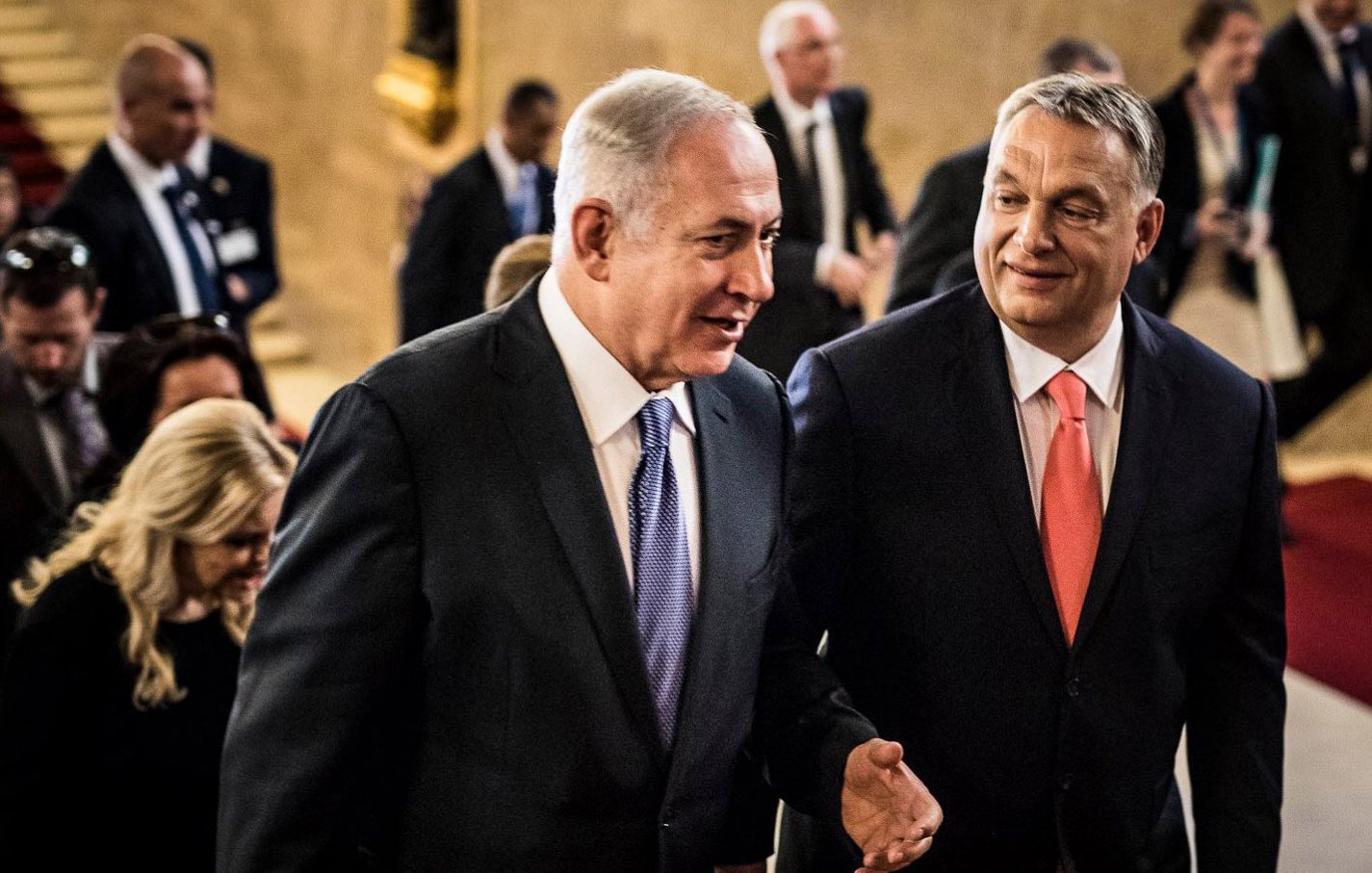 الصورة تُظهر رجلين يتحدثان معًا في ممر يبدو أنه في مبنى رسمي. يظهر أحدهما مرتديًا بدلة داكنة مع رابطة عنق زرقاء، بينما الآخر يرتدي بدلة فاتحة ورابطة عنق برتقالية. خلفهم، يظهر عدد من الأشخاص في بيئة احتفالية. اللغة الجسدية تعكس تفاعلاً ودياً بين الرجلين.