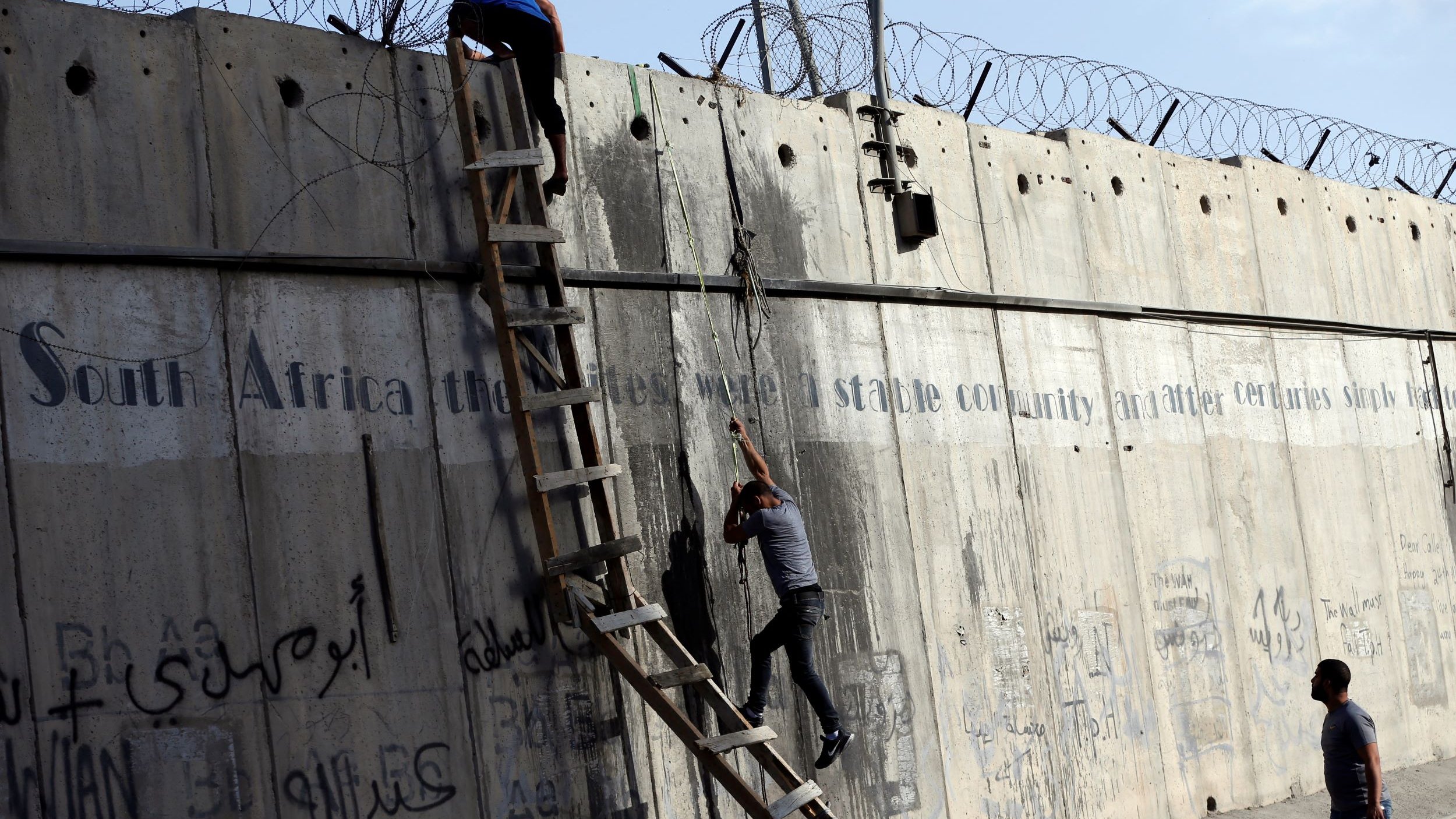 تظهر في الصورة جدارًا خرسانيًا عالياً يؤشر إلى وجود جهد للعبور أو تسلقه. يوجد على الجدار أسلاك شائكة في الأعلى، مما يضيف إلى الأجواء المشدودة. يتسلق أحد الأشخاص الجدار باستخدام سلم خشبي، بينما يحاول الآخر الصعود بشكل متكرر. الجدار مزين بخطوط ورسوم غريبة، بالإضافة إلى كتابات باللغة العربية. تعكس الصورة شعورًا بالجهد والمثابرة في مواجهة عوائق.