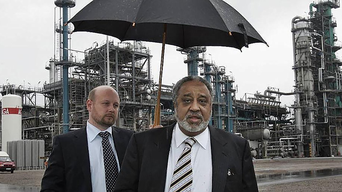 الصورة تظهر رجلين في موقع صناعي. الرجل في المقدمة يحمل مظلة سوداء، ويبدو أنه يتحدث أو يتفاعل مع شخص آخر. الرجل الثاني يقف خلفه بنظرة جدية، ويرتدي بدلة رسمية. في الخلفية، توجد منشآت صناعية تظهر أنابيب ومعدات، مما يشير إلى أن المكان هو مصنع أو منشأة صناعية. السماء غائمة، مما يدل على أنه قد يكون هناك مطر.