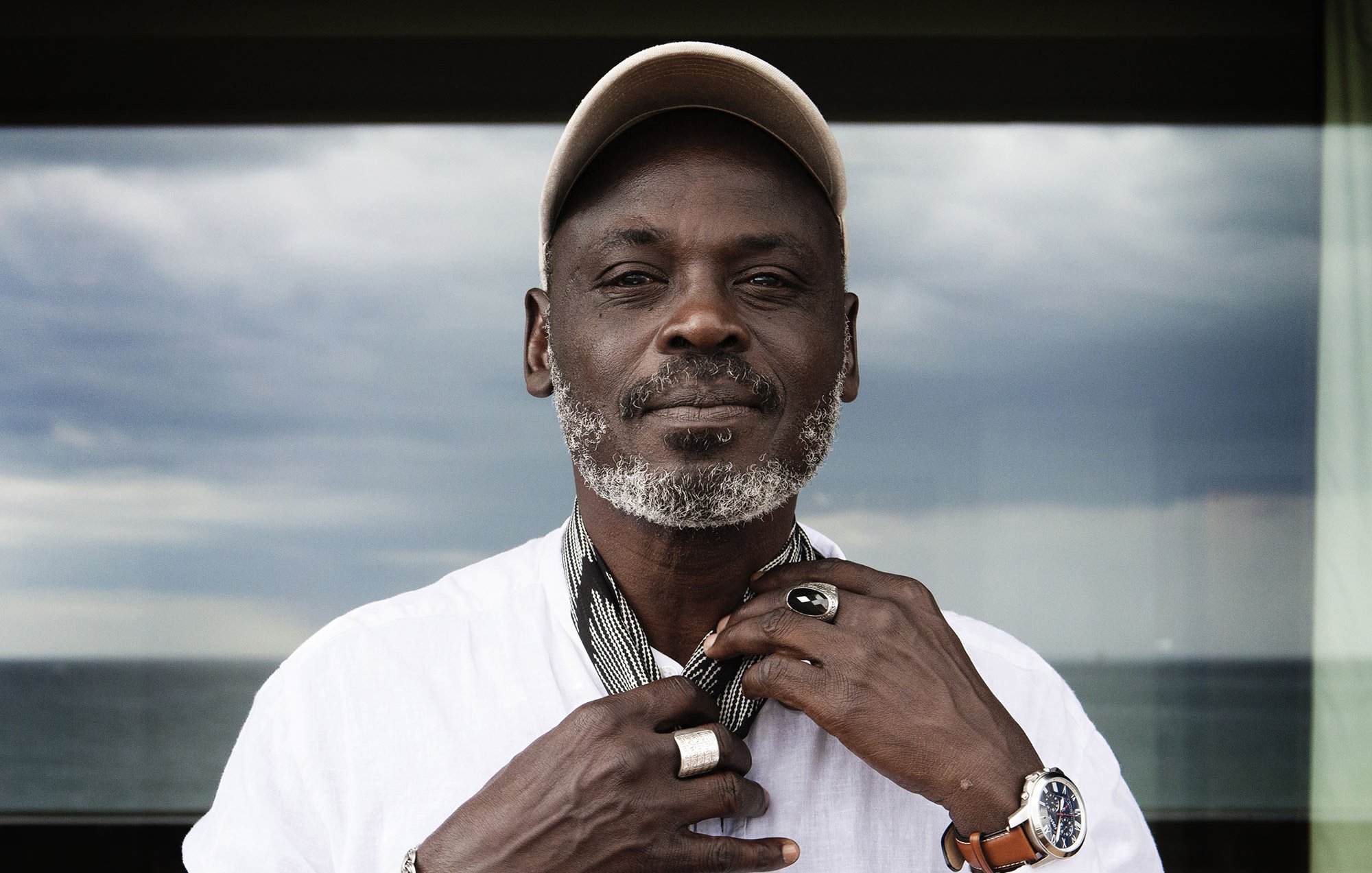 تظهر الصورة رجلًا يرتدي قميصًا أبيض وسلسلة ويضع قبعة على رأسه. يبدو أنه يثبت وشاحًا حول رقبته، ويقف أمام منظر طبيعي يضم البحر في الخلفية. السماء تظهر غائمة وكأنها تحمل بعض الغيوم. ملامح وجهه تعكس الثقة والهدوء.