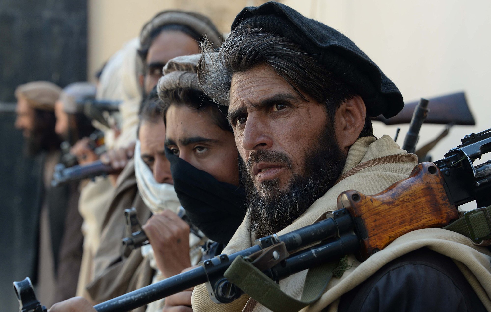 تظهر الصورة مجموعة من الرجال يقفون في صف، يحمل كل منهم سلاحًا. معظمهم يرتدي ملابس تقليدية، ويبدو عليهم الجدية والتركيز. يتواجد في الخلفية مجموعة من الأشخاص أيضًا، ويبدو أن الصورة تلتقط لحظة توتر أو استعداد. الخلفية غير واضحة ولا تحتوي على تفاصيل كثيرة.