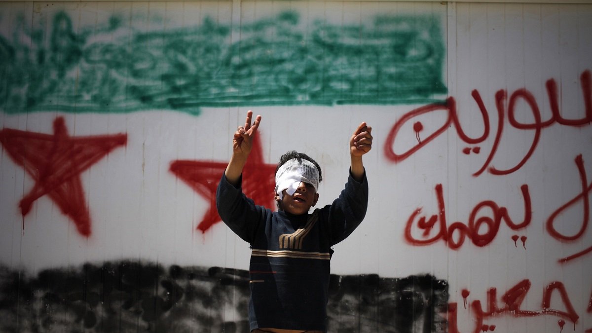 تظهر الصورة صبيًا صغيرًا يرفع يديه بإشارات النصر، بينما يغطّي عينيه برباط أبيض. خلفه جدار مطلي عليه رسم يمثل علم سوريا، مع كلمات مكتوبة بالعربية تظهر اعتراضًا أو دعماً لموضوع معين. تعكس الصورة مشاعر الأمل والعزيمة في وجه الصعوبات.