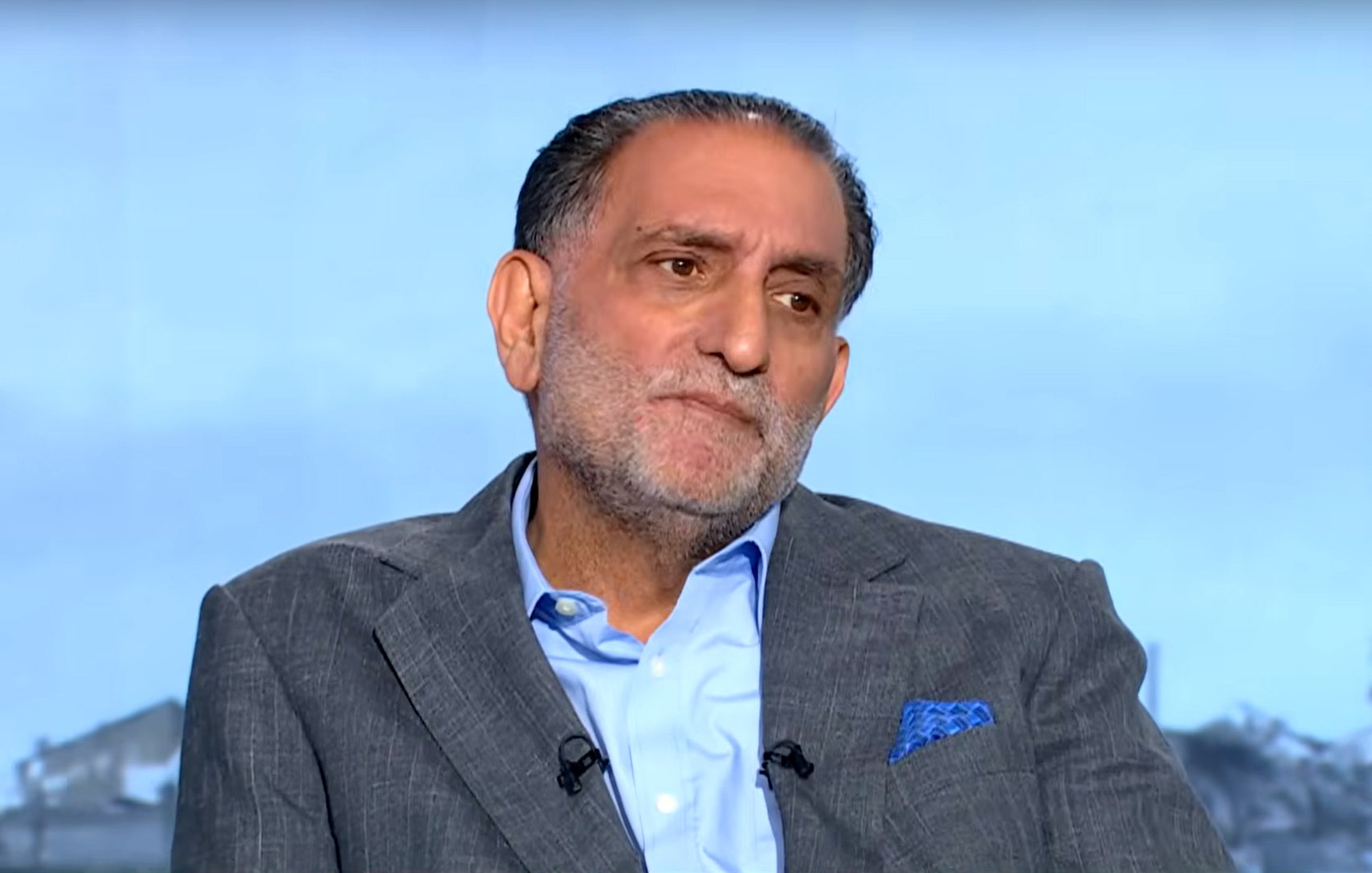 رجل يجلس مرتديًا بدلة رمادية، يبدو عليه التفكير والاهتمام.