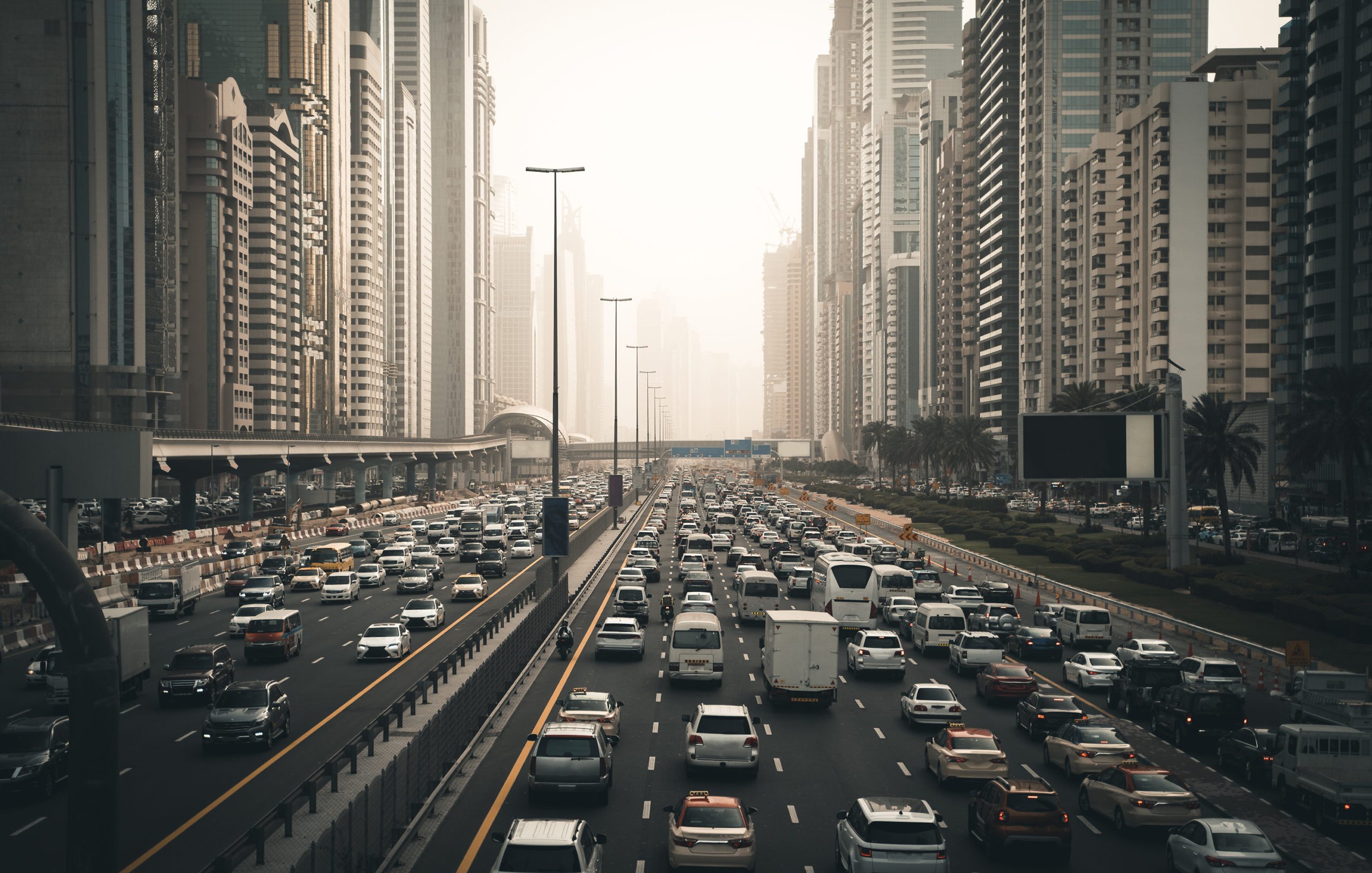 تظهر في الصورة مشهدًا حضريًا لمدينة مزدحمة، حيث تتواجد الكثير من السيارات المتوقفة في زحام مروري على عدة مسارات. تحيط بالمكان ناطحات السحاب العالية التي تدل على التطور العمراني في المنطقة. تبدو السماء مغشاة بضباب خفيف، مما يعطي انطباعًا عن أجواء المدينة في وقت متأخر من اليوم أو خلال الصباح الباكر. هناك مساحات من الأشجار على جوانب الطريق تضفي لمسة من الطبيعة وسط الحضر.