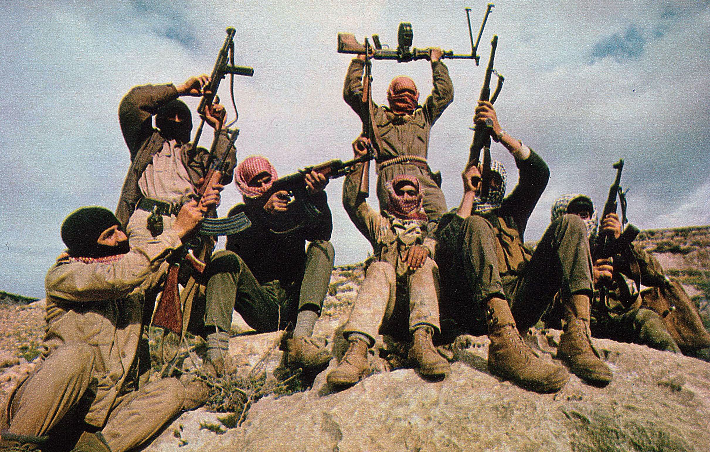 الصورة تظهر مجموعة من الأشخاص يرتدون ملابس عسكرية، وهم يتجهون نحو الكاميرا. بعضهم يحمل بنادق، بينما يقوم شخص في المنتصف برفع سلاح فوق رأسه. يظهر الأشخاص في وضعية متحمسة على صخرة كبيرة، وتظهر الخلفية سماء زرقاء مع بعض الغيوم. تعبّر الصورة عن روح الرفاقية والتصميم.