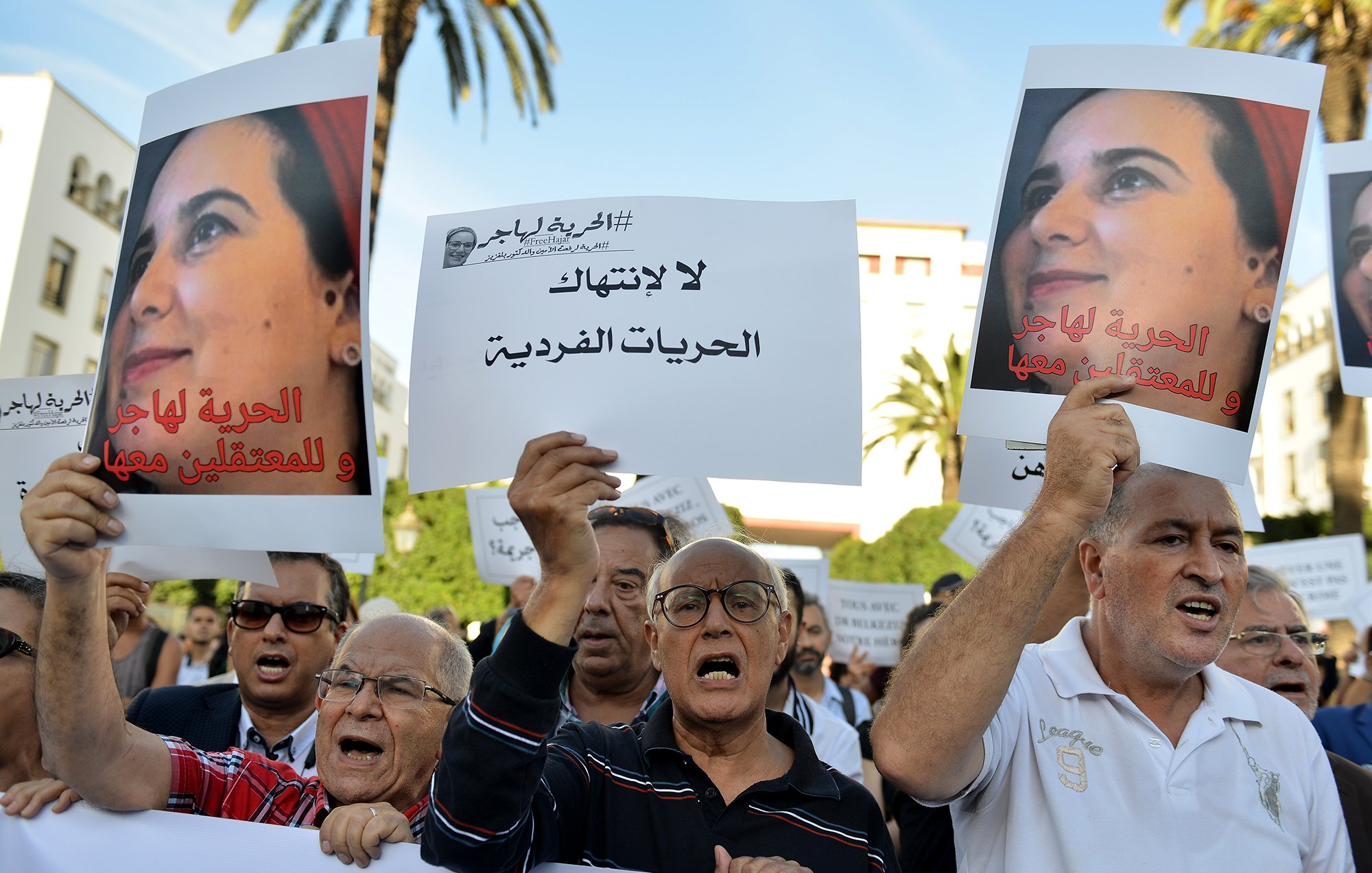 تظهر في الصورة مجموعة من الناس يتظاهرون في مكان عام، حاملين لافتات مكتوبة. يبدو أنهم يعبرون عن مطالب تتعلق بالحرية الفردية وحقوق المعتقلين. المشهد يدل على حماس المشاركين، حيث يعبر بعضهم عن استيائهم بصوت عالٍ. في الخلفية، يمكن رؤية بعض النخيل والسماء التي تحتوي على بعض السحب، مما يدل على أن المظاهرة جرت في يوم مشمس.