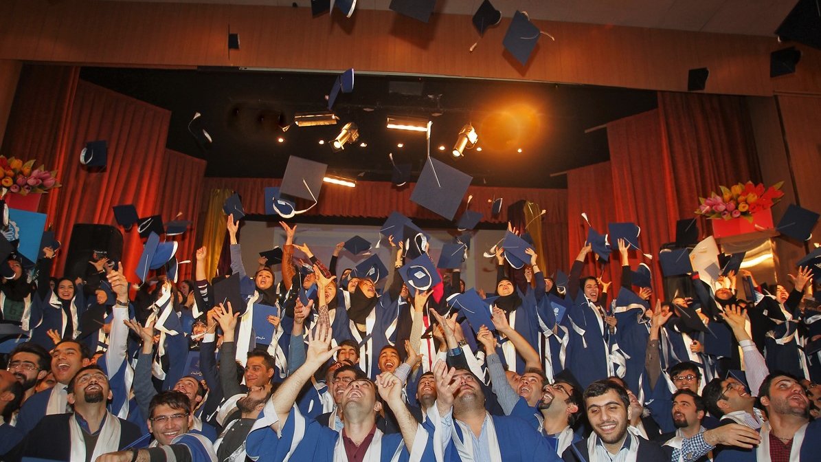 تظهر الصورة مجموعة كبيرة من الطلاب في حفل تخرج، وهم يرتدون قبعات التخرج والعباءات التقليدية. يبدو الجميع في حالة من الفرح والسعادة، حيث يرفعون قبعاتهم في الهواء احتفالاً بالإنجاز. الخلفية مليئة بالزينة، وهناك أضواء مسلطة تعزز من أجواء الحفل. تعكس الصورة روح الفخر والنجاح في هذه المناسبة المهمة في حياة الطلاب.