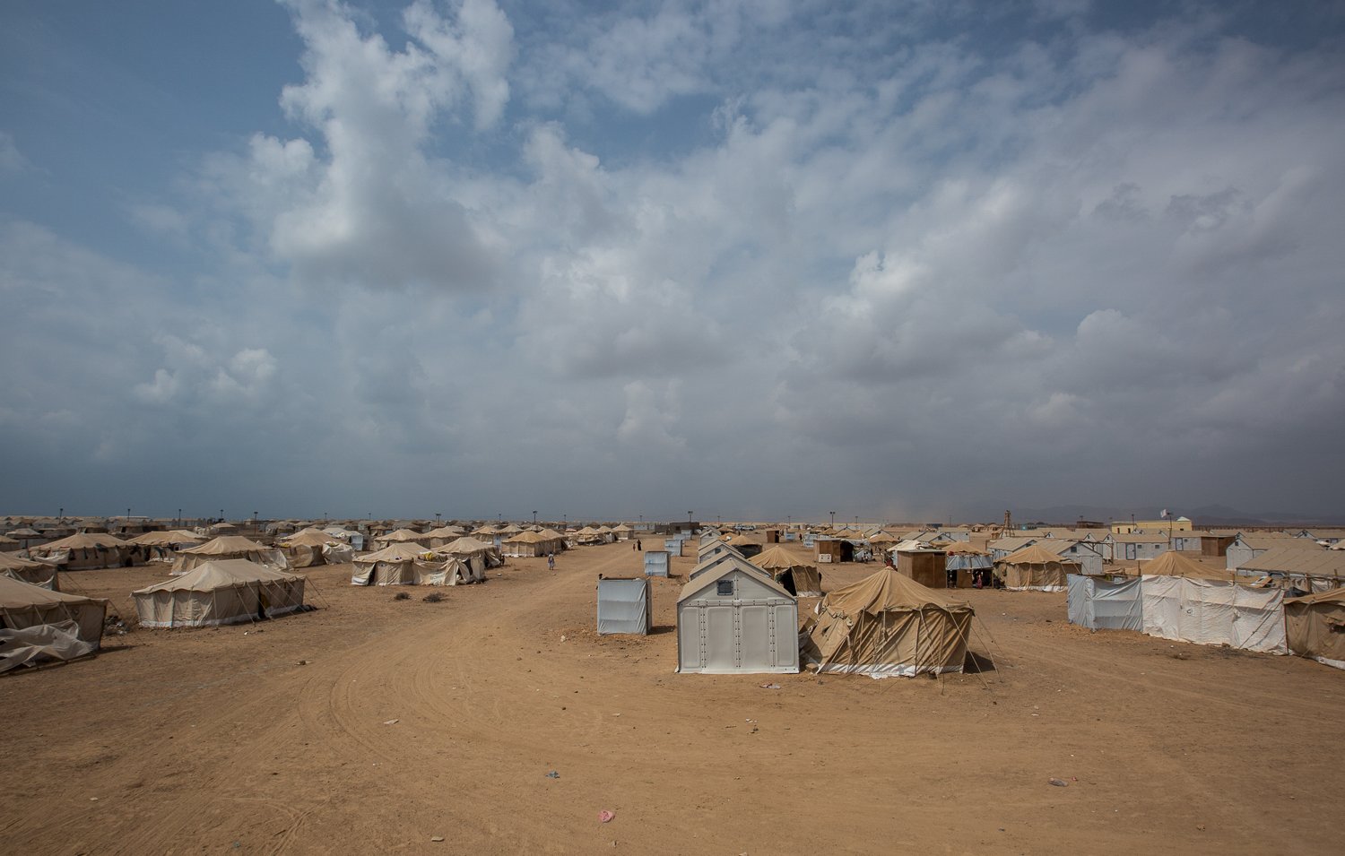 تظهر في الصورة مشهدًا صحراويًا واسعًا، حيث تنتشر العديد من الخيام على الأرض الرملية. السماء ملبدة بالسحب، مما يوحي بوجود طقس متغير. يبدو أن المكان يحمل طابعًا يجمع بين البساطة والاحتياجات الأساسية، حيث تُستخدم الخيام كوسيلة للإيواء. تبرز بعض الخيام بألوان فاتحة ومختلفة، مما يضفي طابعًا متنوعًا على المنطقة.