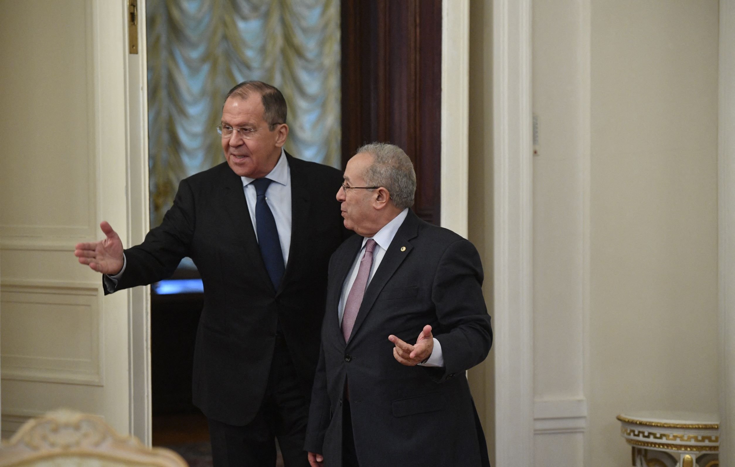 الصورة تظهر شخصين في غرفة داخلية ذات تصميم كلاسيكي. يبدو أن أحدهم يتحدث ويشير بيده، بينما الآخر يستمع. الإضاءة في الغرفة دافئة، وهناك ستائر ونوافذ خلفهم. الملابس الرسمية تشير إلى أن اللقاء قد يكون ذو طابع رسمي أو دبلوماسي.