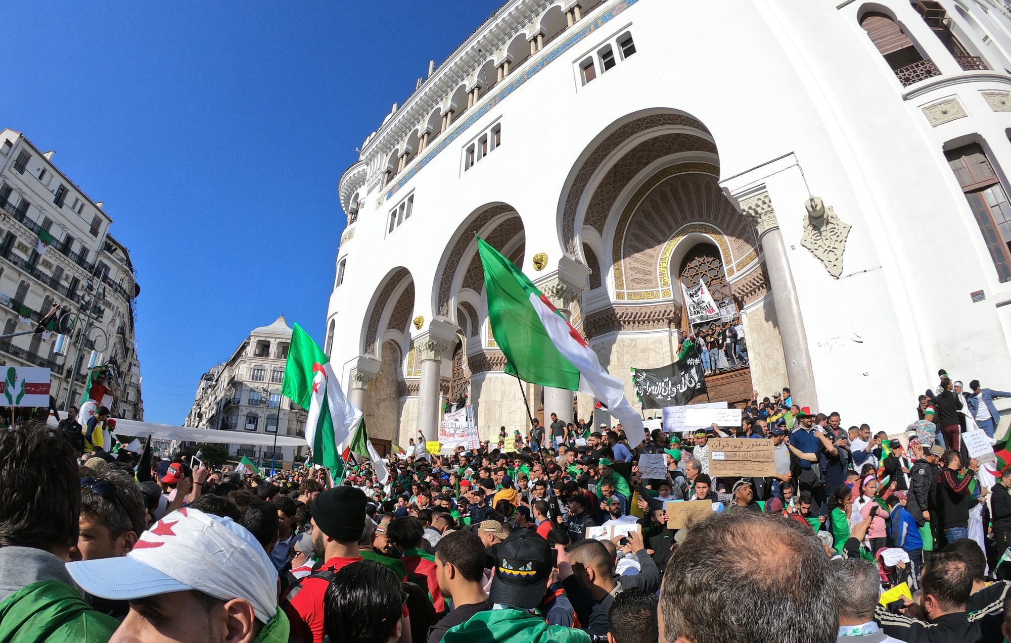 تظهر الصورة تجمعًا حاشدًا لمتظاهرين في مكان مفتوح، يحتشدون أمام مبنى تاريخي. يرفع المتظاهرون أعلام الجزائر بينما يحمل البعض لافتات تحمل شعارات. يتواجد عدد كبير من الأشخاص في الصورة، مما يبرز حماسهم ومشاركتهم الفاعلة. السماء زرقاء وظهور شعاع الشمس، مما يعكس أجواء نابضة بالحياة.
